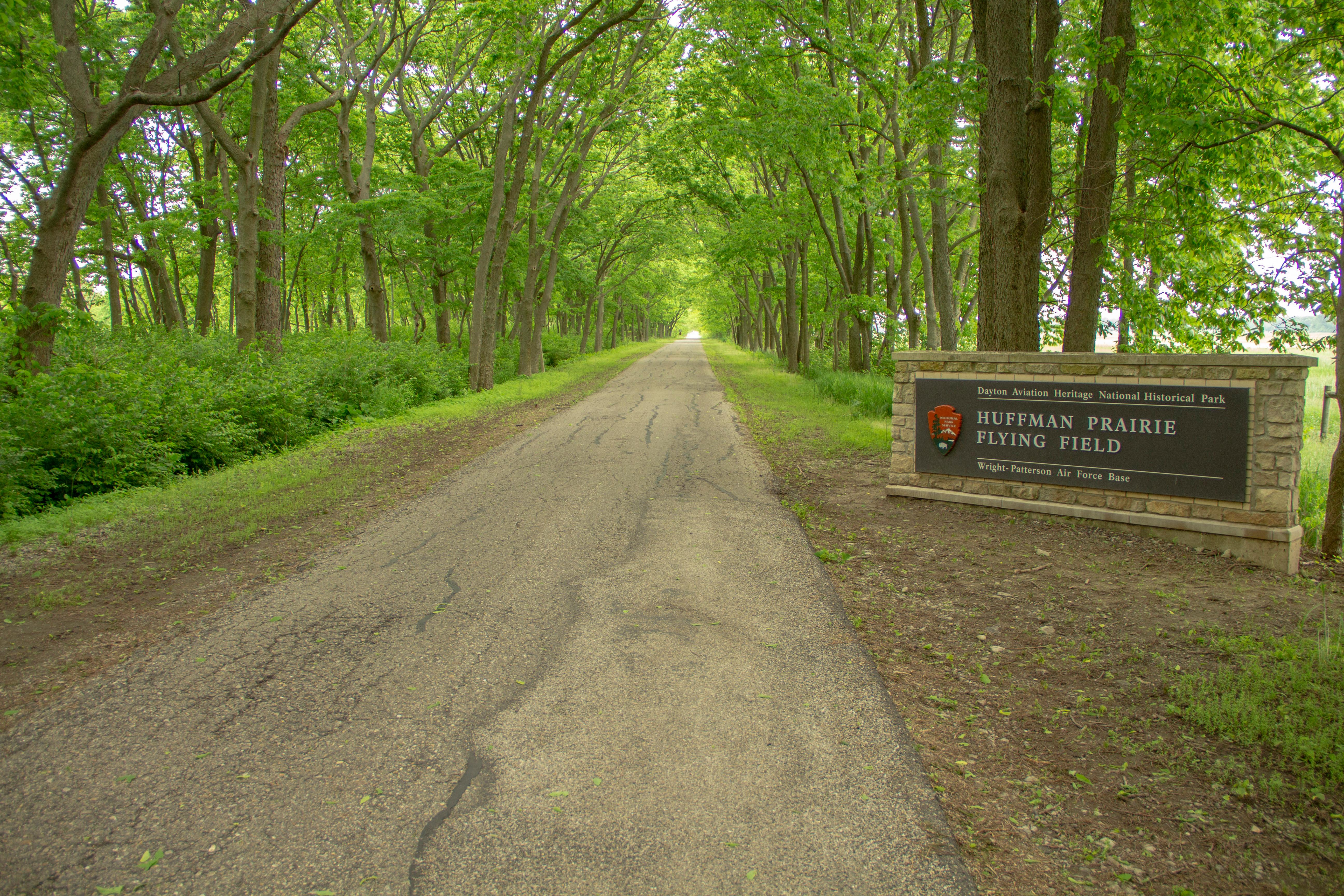 A dirt road between tall trees with a stone sign reading Huffman Prairie Flying Field.