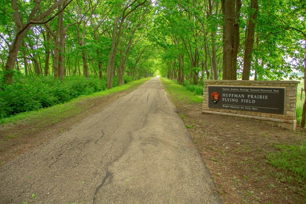 A dirt road between tall trees with a stone sign reading Huffman Prairie Flying Field.