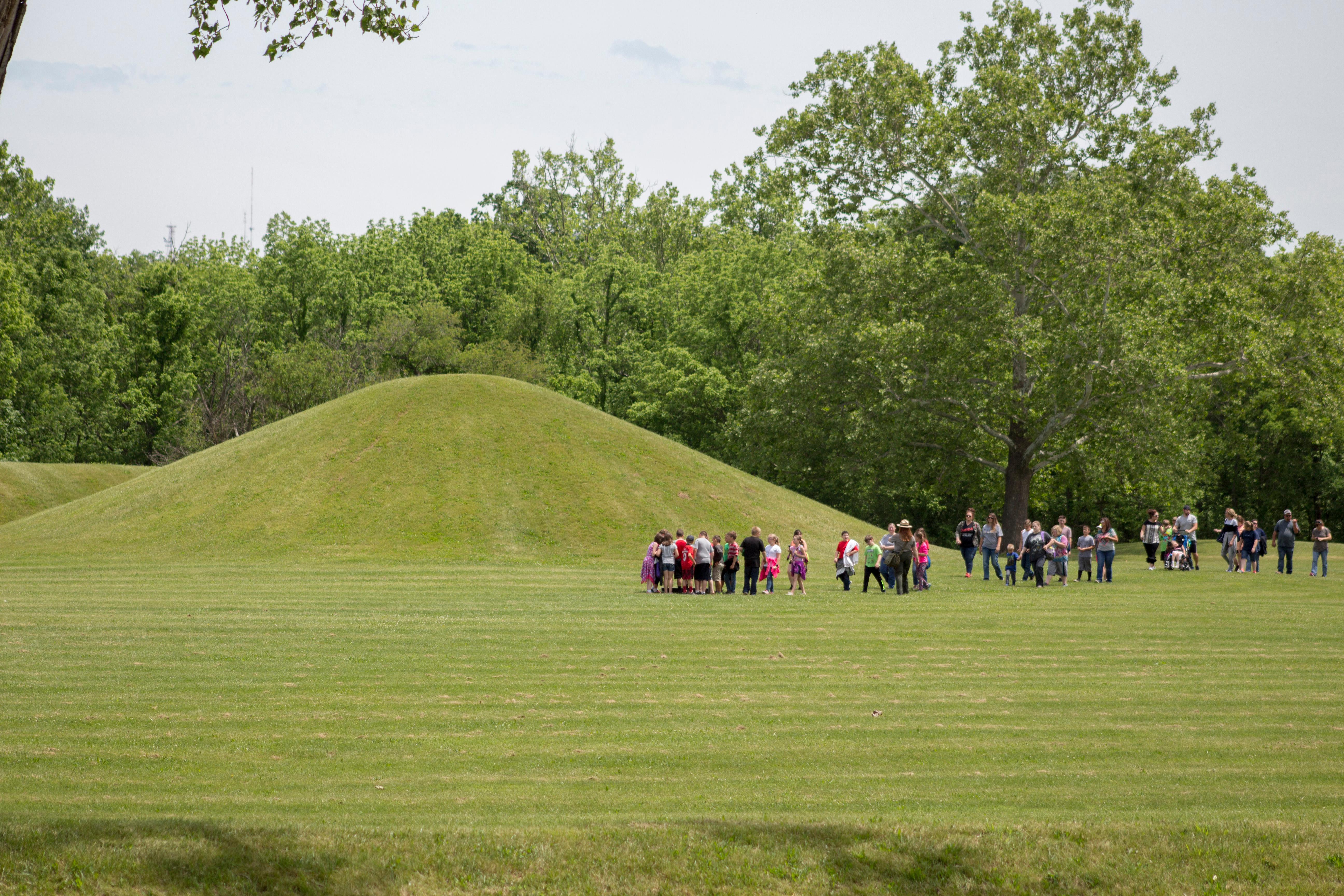 Several children walking with a park ranger in a flat hat near grass-covered mounds.