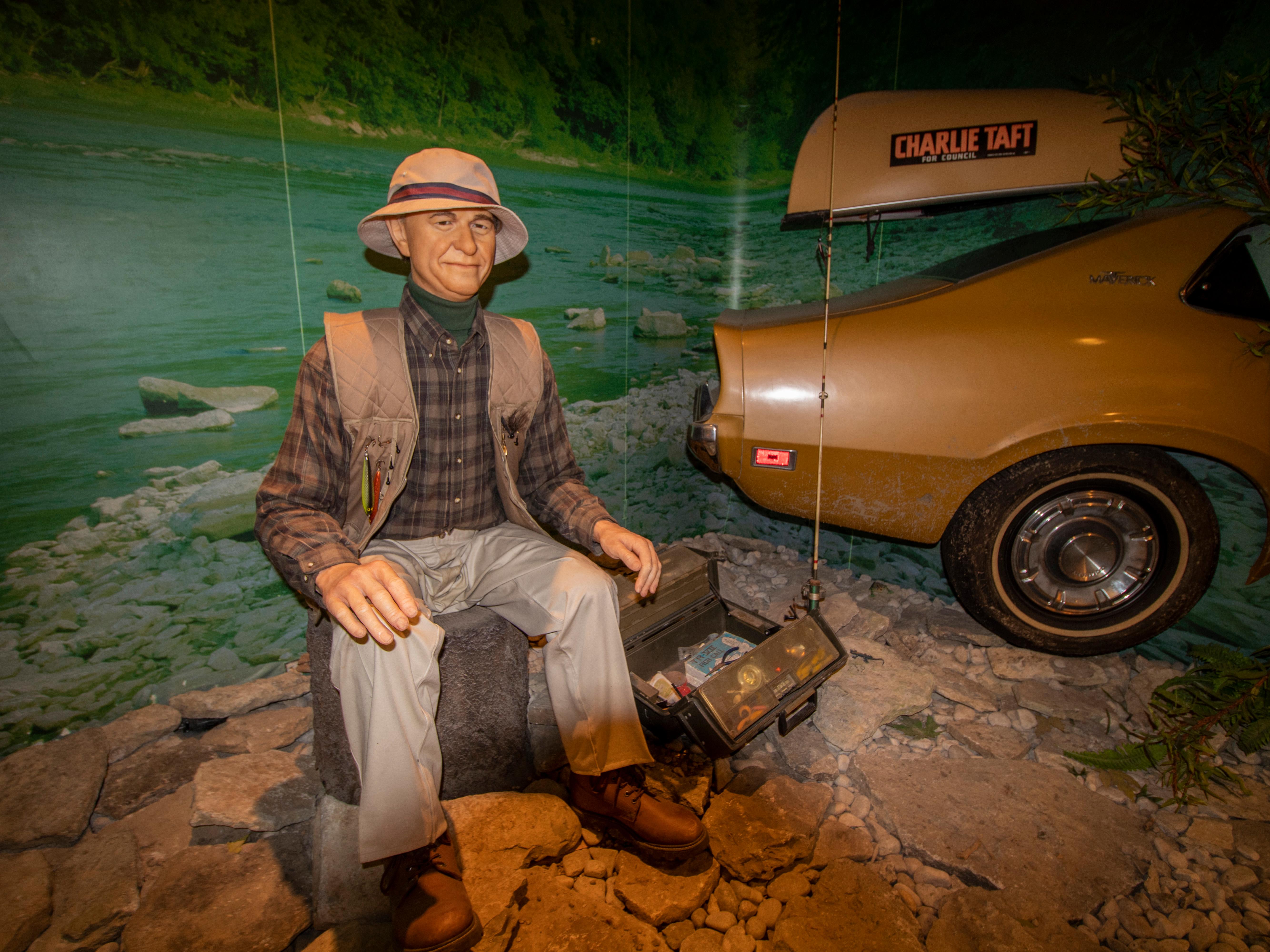 A sitting model of an older man in a vest, shirt and pants next to a replica of a car & canoe on top
