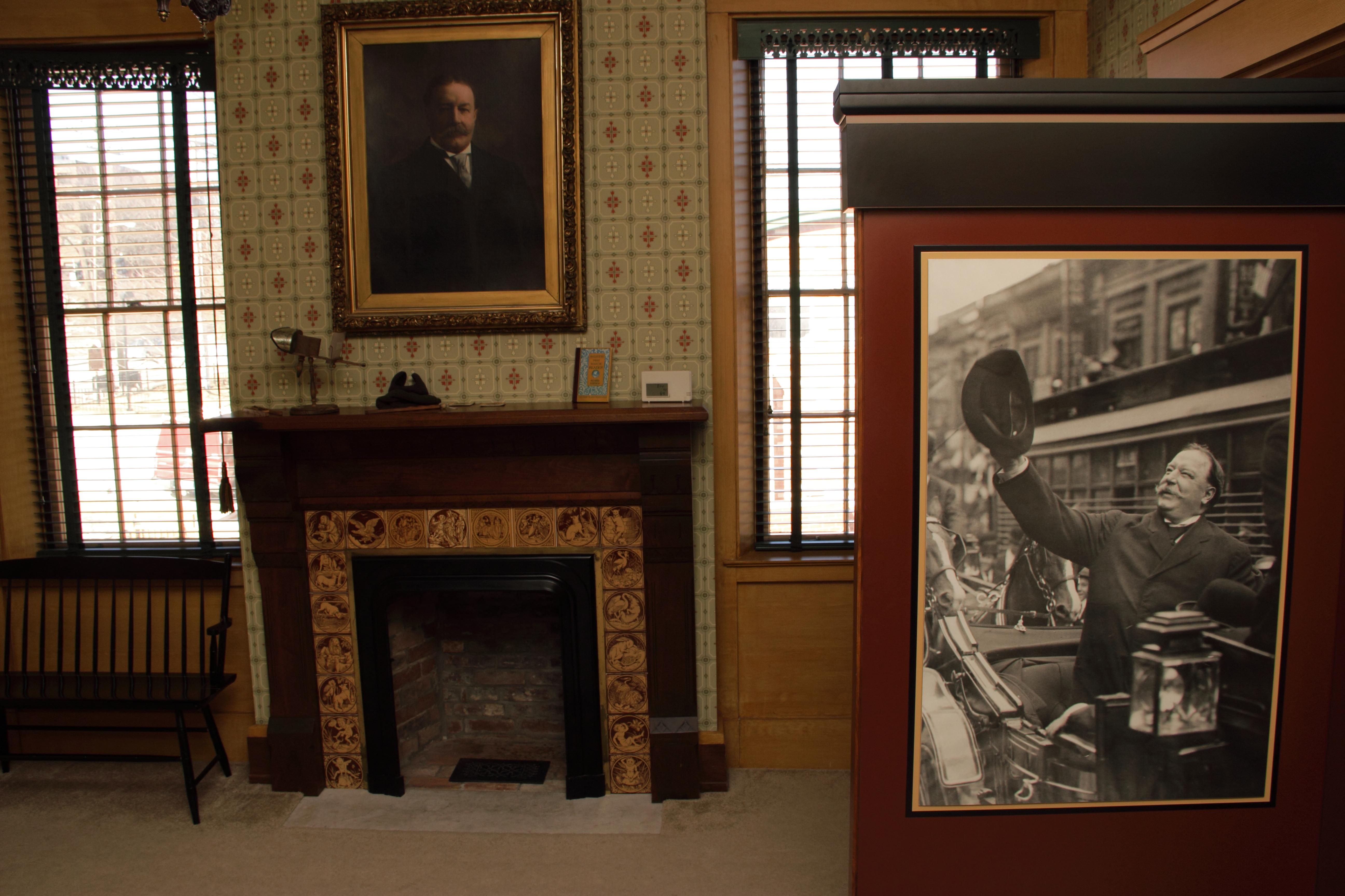 A room inside of a house showing two portraits of men and a fireplace on a wall between two windows.