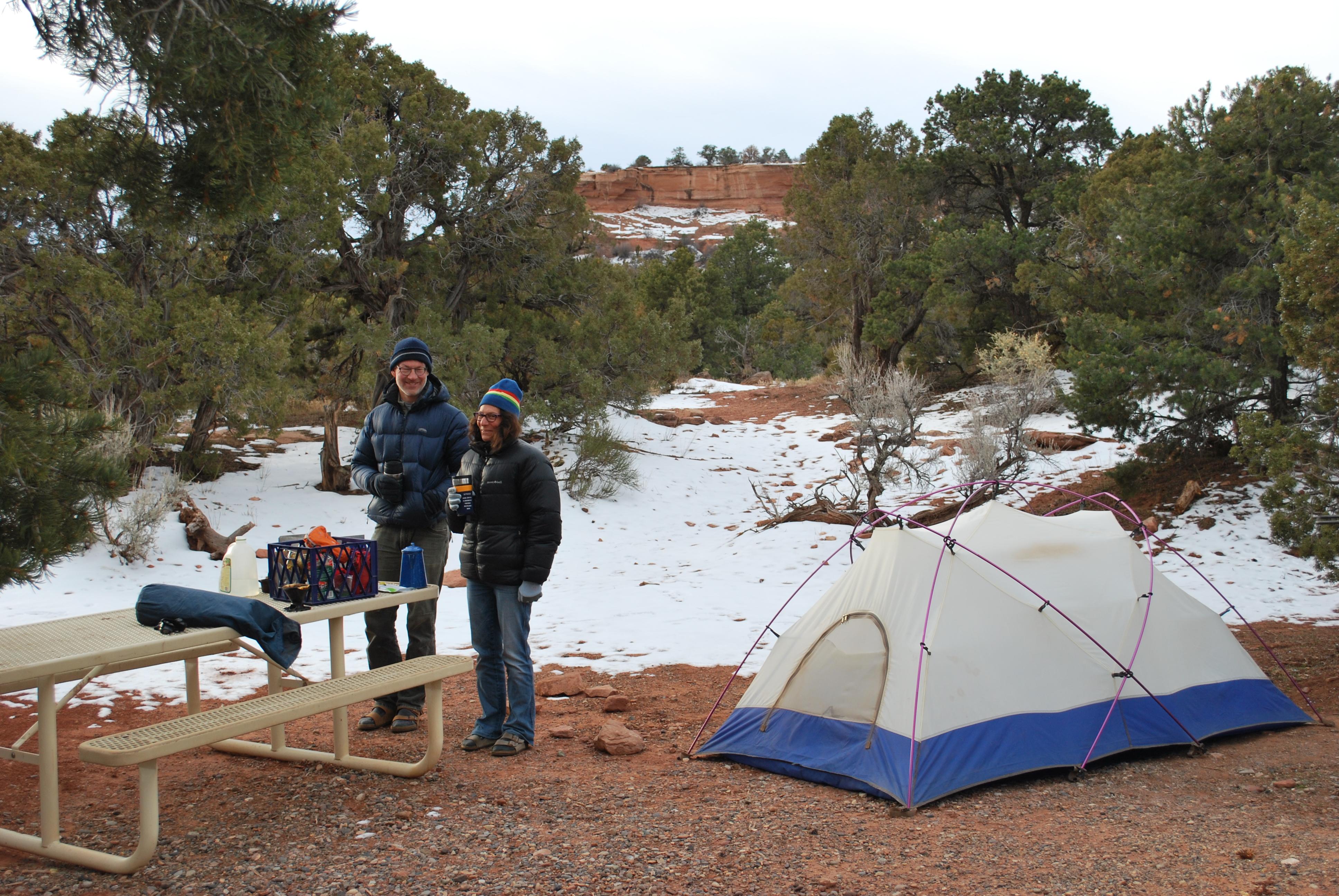 Two people wearing jackets and hats near tent with snow behind tent