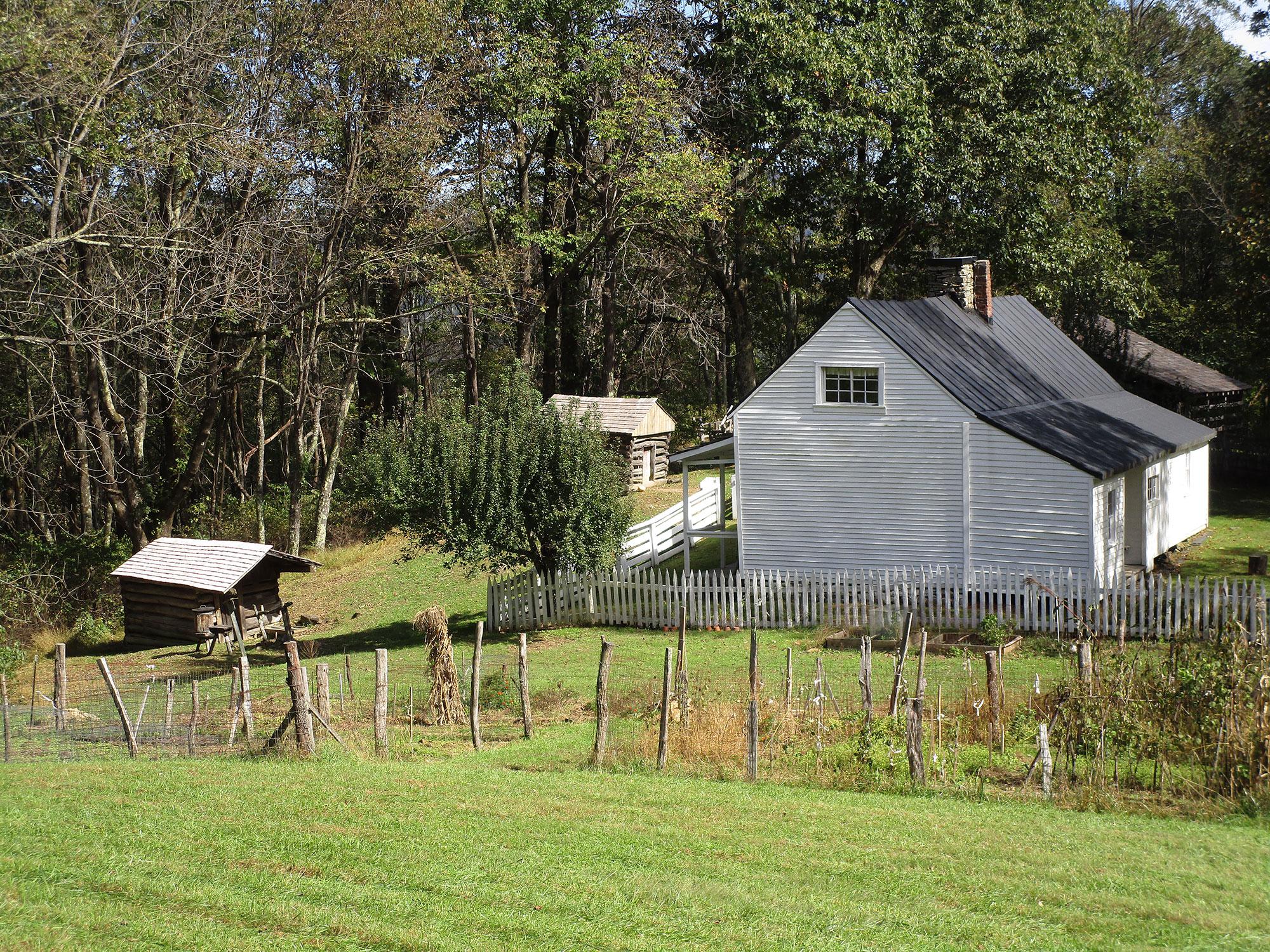 A white, wooden farmhouse surrounded by lawn and fenced garden