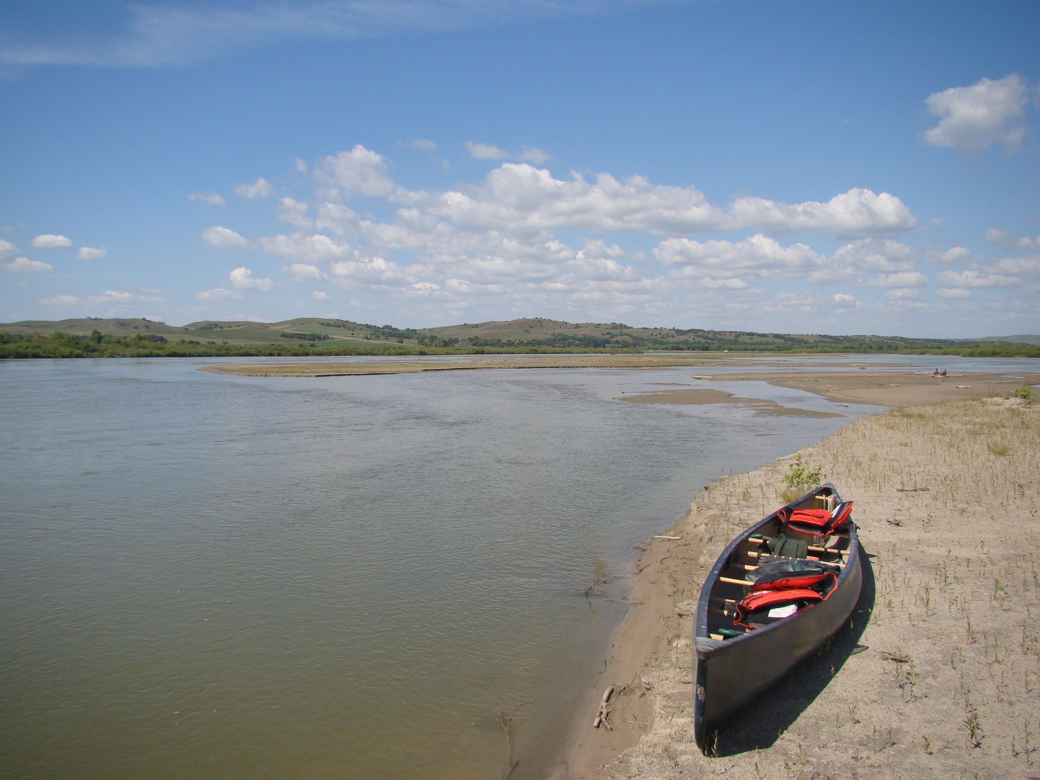 A canoe loaded with gear sits quietly on a sandy river bank with blue sky above.