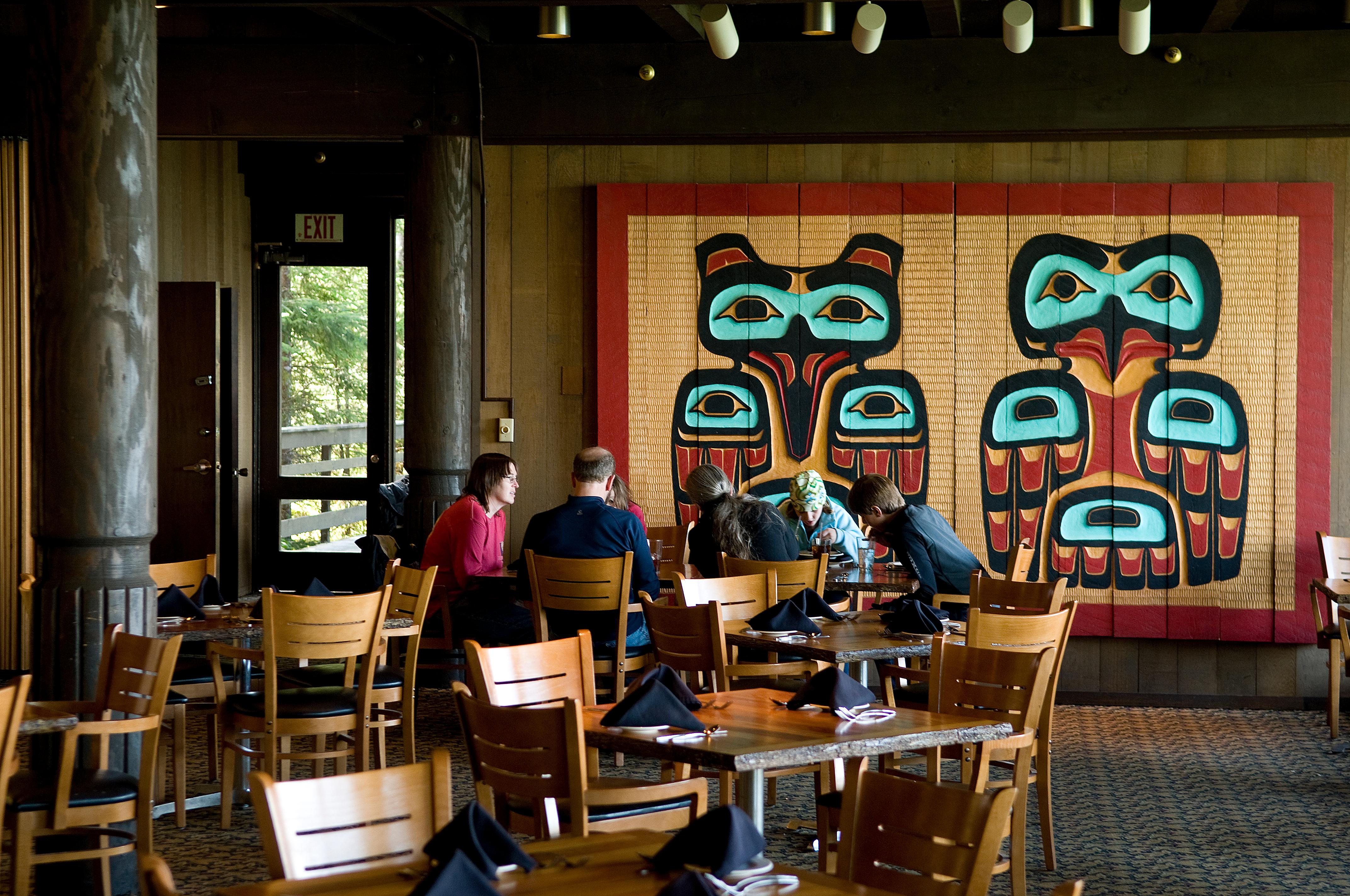 Several people sit at a dining table in front of Tlingit form line wall art inside Glacier Bay Lodge