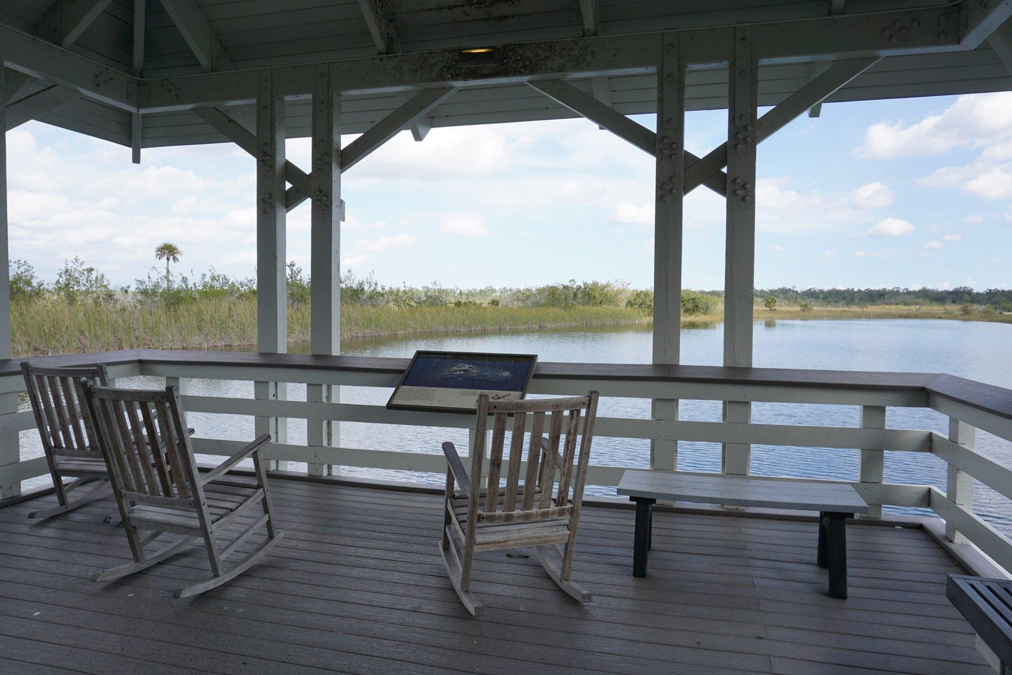 Rocking chairs sit lazily in the shade on a wooden platform overlooking a small lake