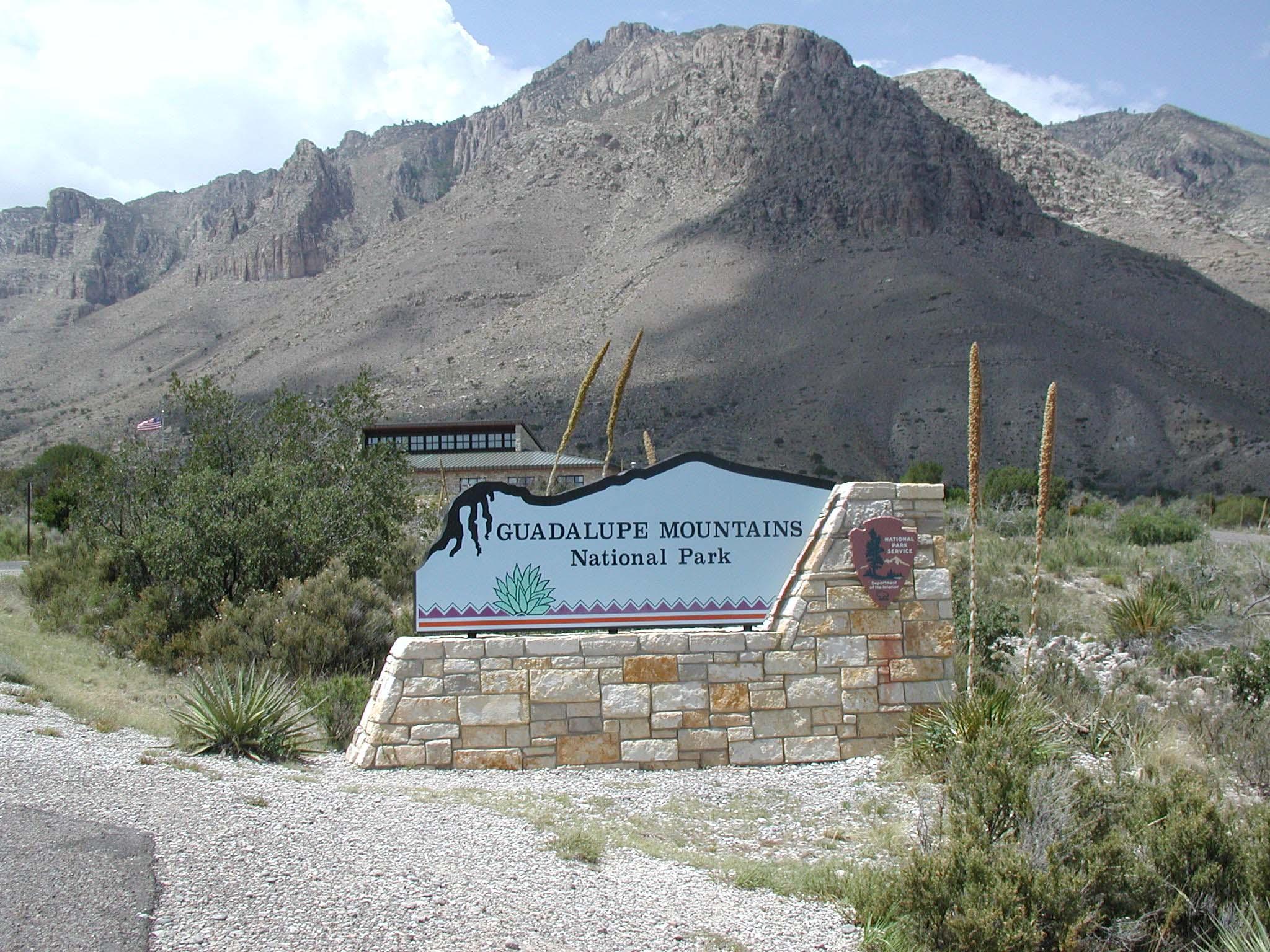 Behind the park entry sign is the roof line of a visitor center in front of Hunter Peak