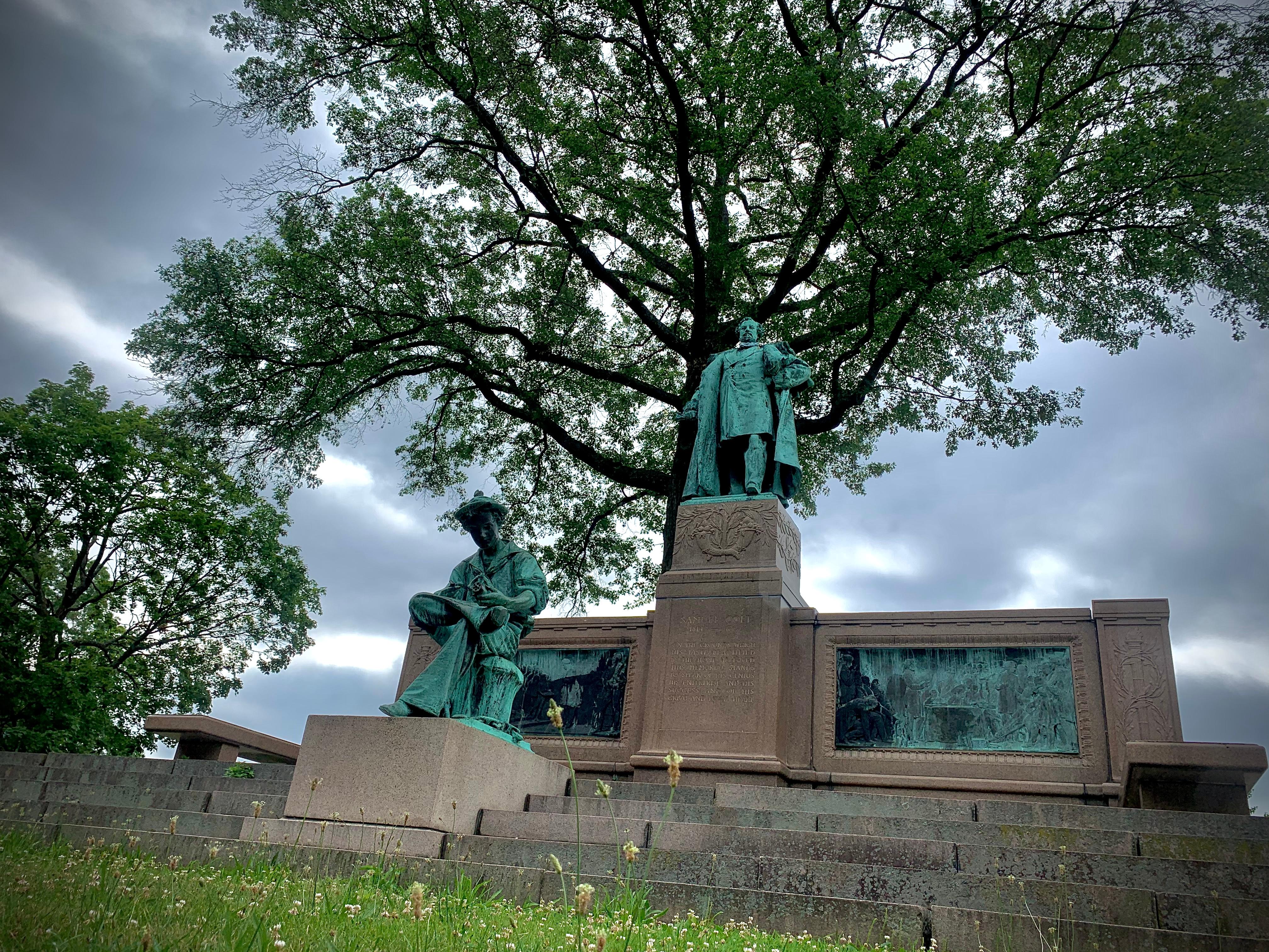 A statue of a young and an adult Samuel Colt in front of a tree with a cloudy sky.