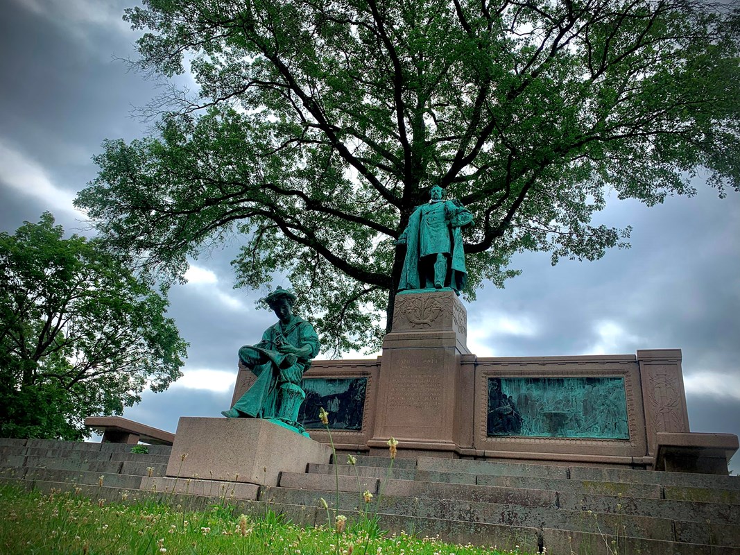 A statue of a young and an adult Samuel Colt in front of a tree with a cloudy sky. 