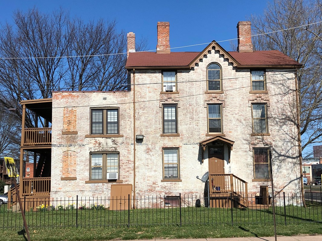 A white stone building with multiple windows and three chimneys with green grass and a black fence. 