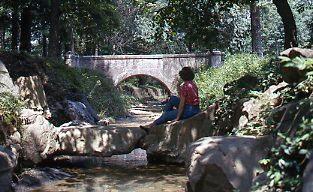Hiker sits at the edge of the creek, with a bridge in the background