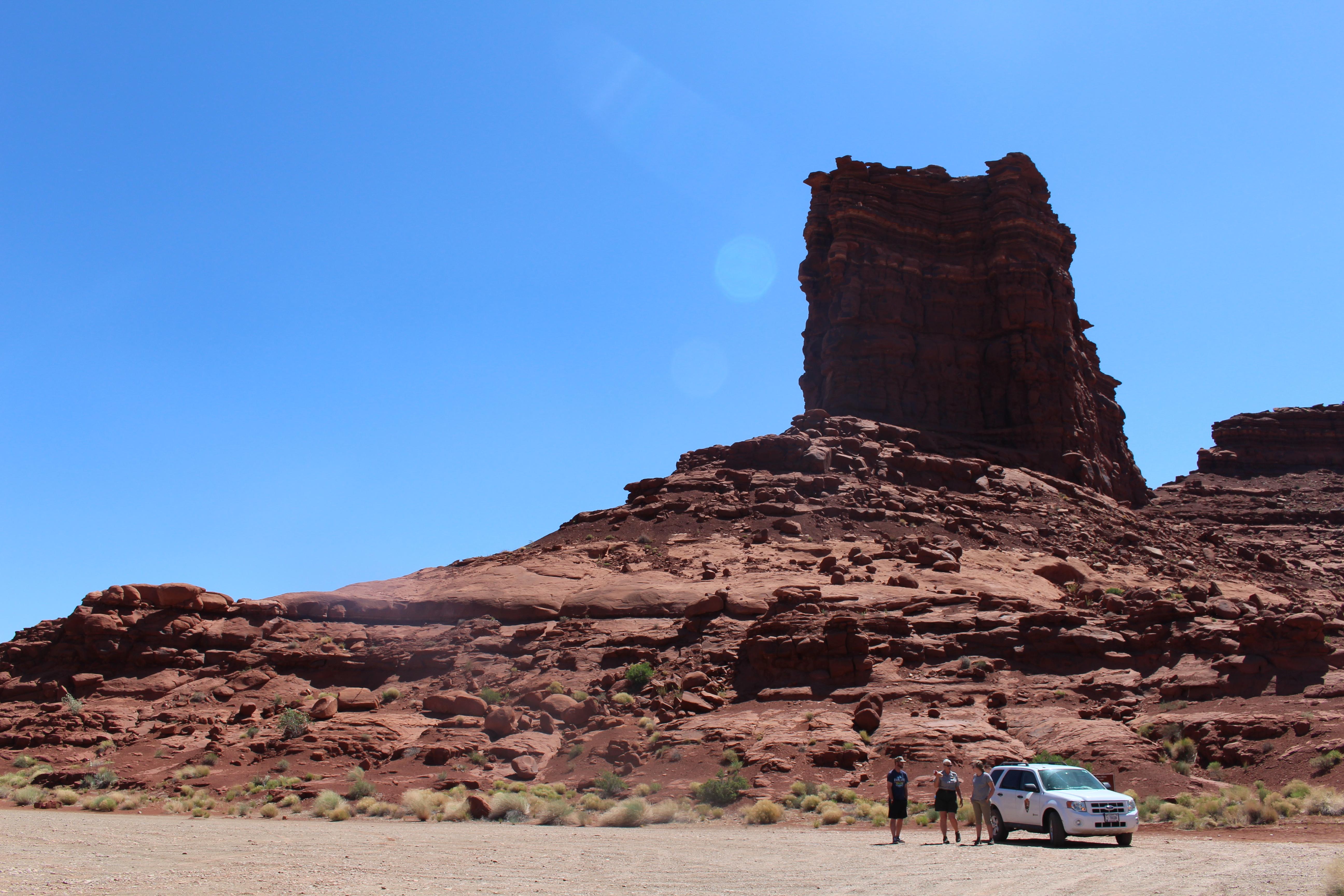 Three people and a vehicle at the base of a large sandstone butte