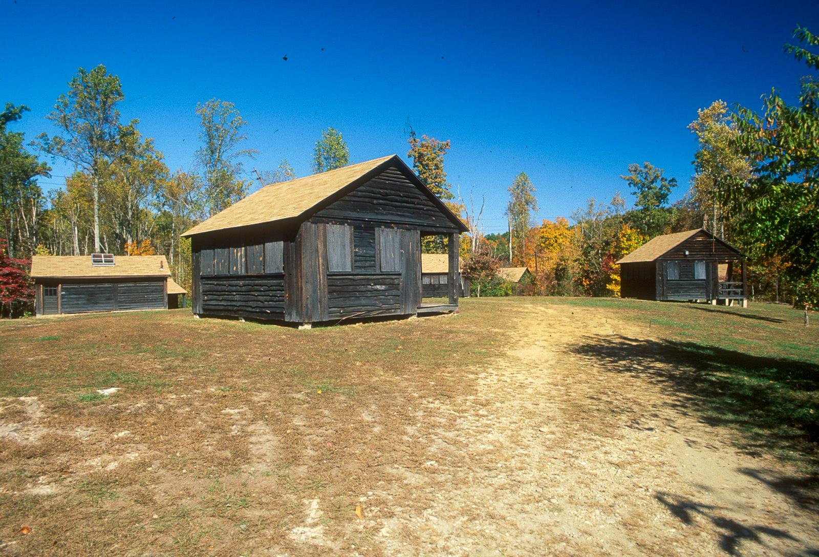 A wooden, dark brown sleeping cabin sits in a field. Behind it are other cabins and trees.