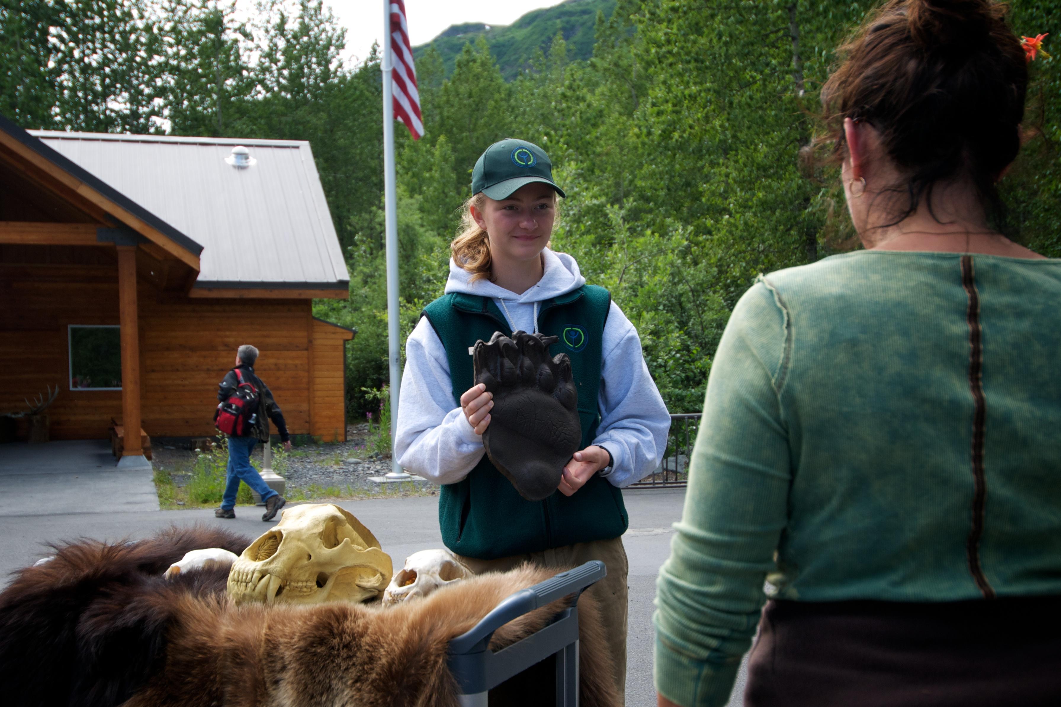 A youth park staff talks to visitors with a bear print mold in hand next to a bear skull on pelt.