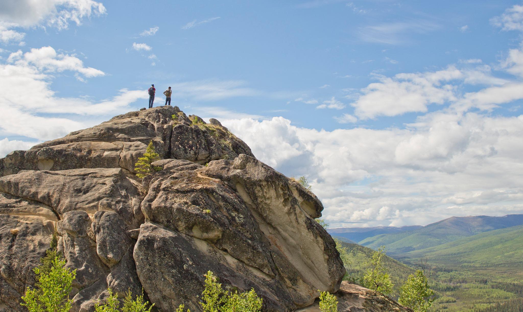 Two Hikers stand on top of a very large granite boulder, looking out across the landscape