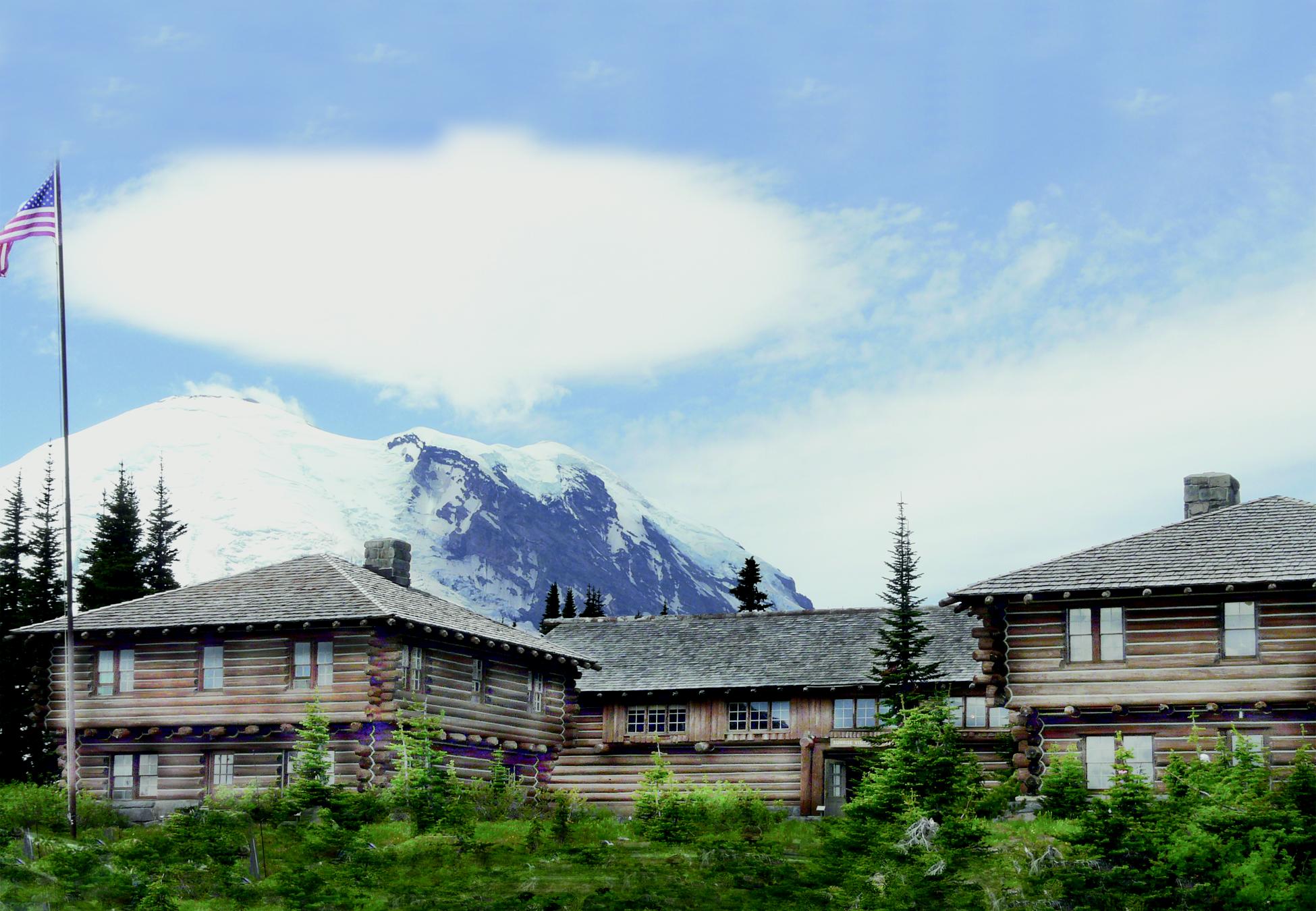 An American flag waves in front of 3 rustic wooden buildings with the peak of Mount Rainier behind.