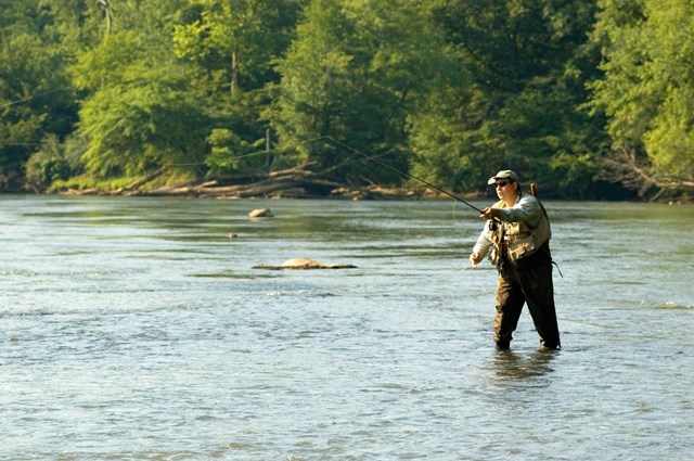 Angler in waders holding a fishing rod in the middle of a shallow river