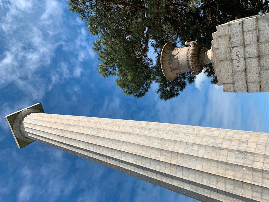 352 foot tall stone memorial column rises up to touch blue sky. In front a 5' urn on pedestal