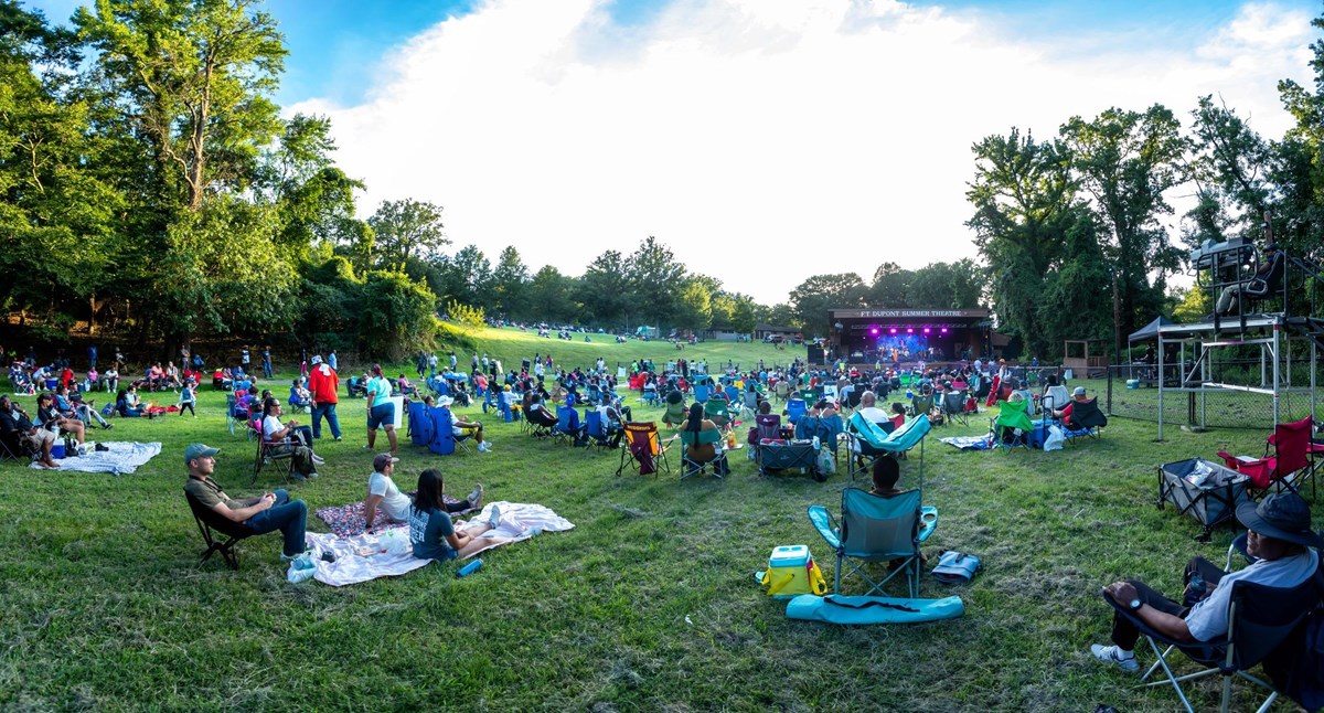 Large crowd sitting on blankets and in folding chairs in front of an outdoor stage