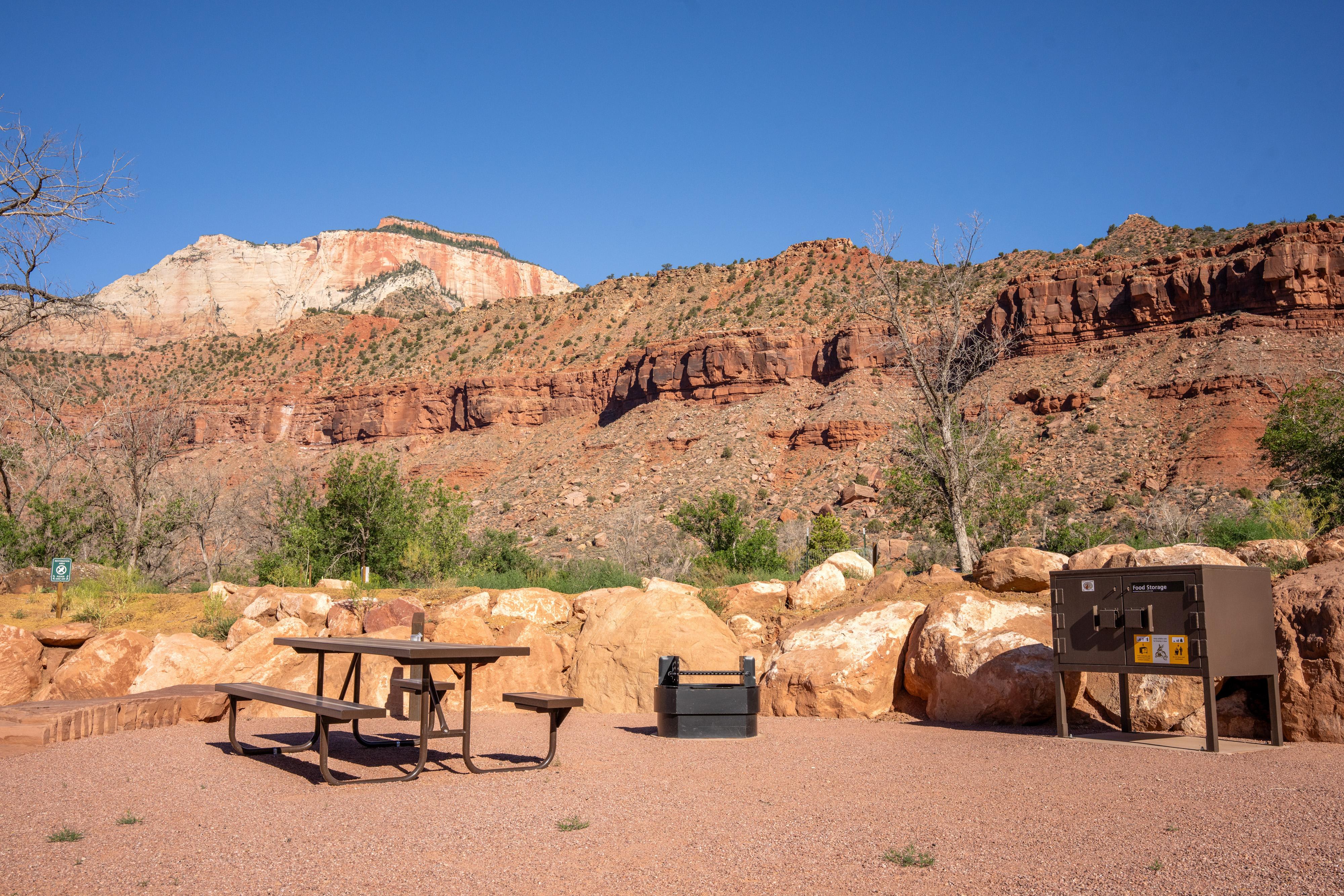 A gravel campsite lined with boulders. A picnic table, fire ring, and storage box sit in the site.