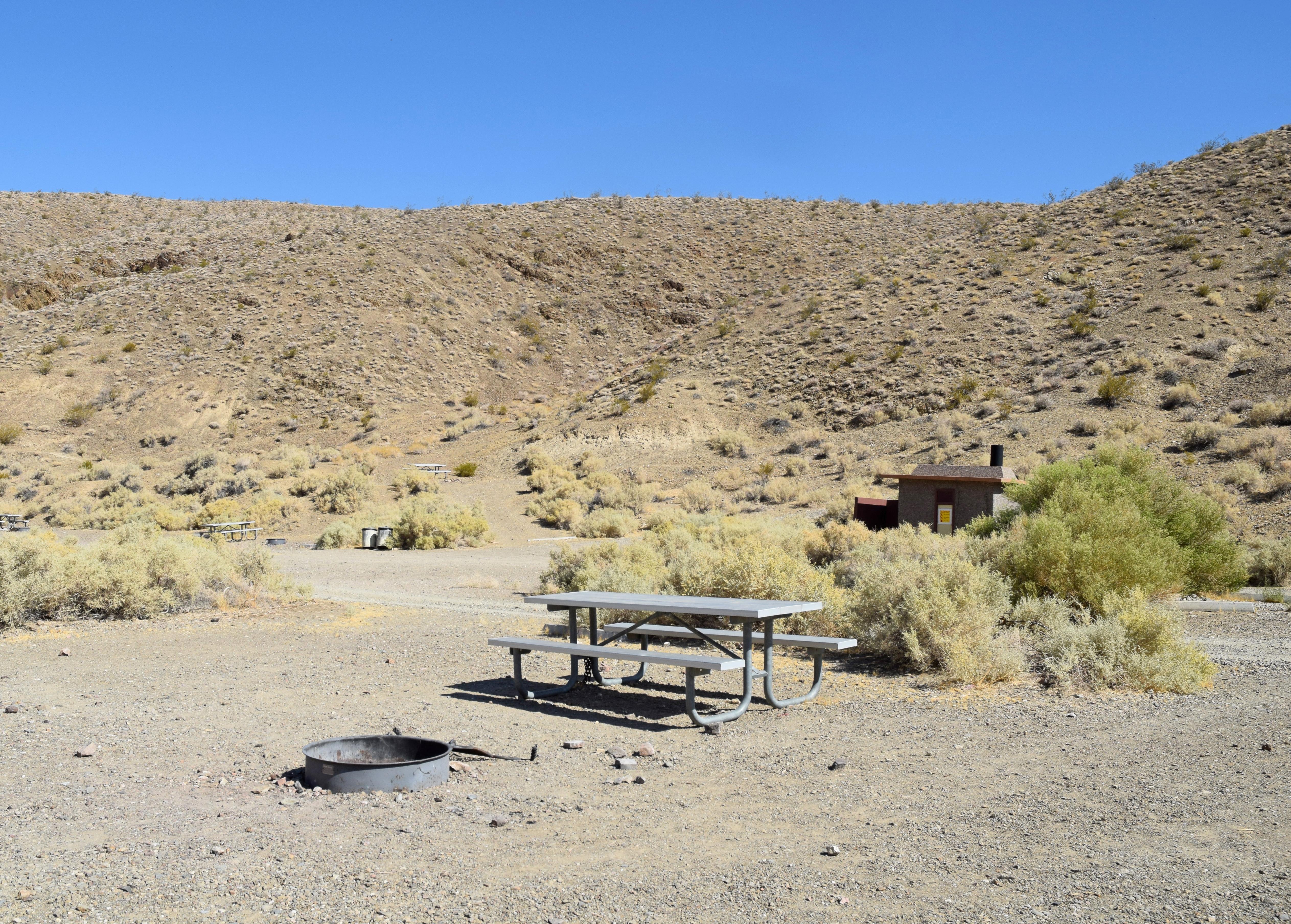Large flat, packed dirt & gravel area with dispersed small bushes. One hill covers background.