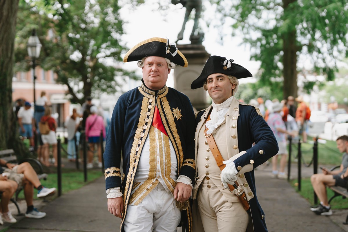 Two reenactors in Revolutionary War-era military uniforms stand together in a park.