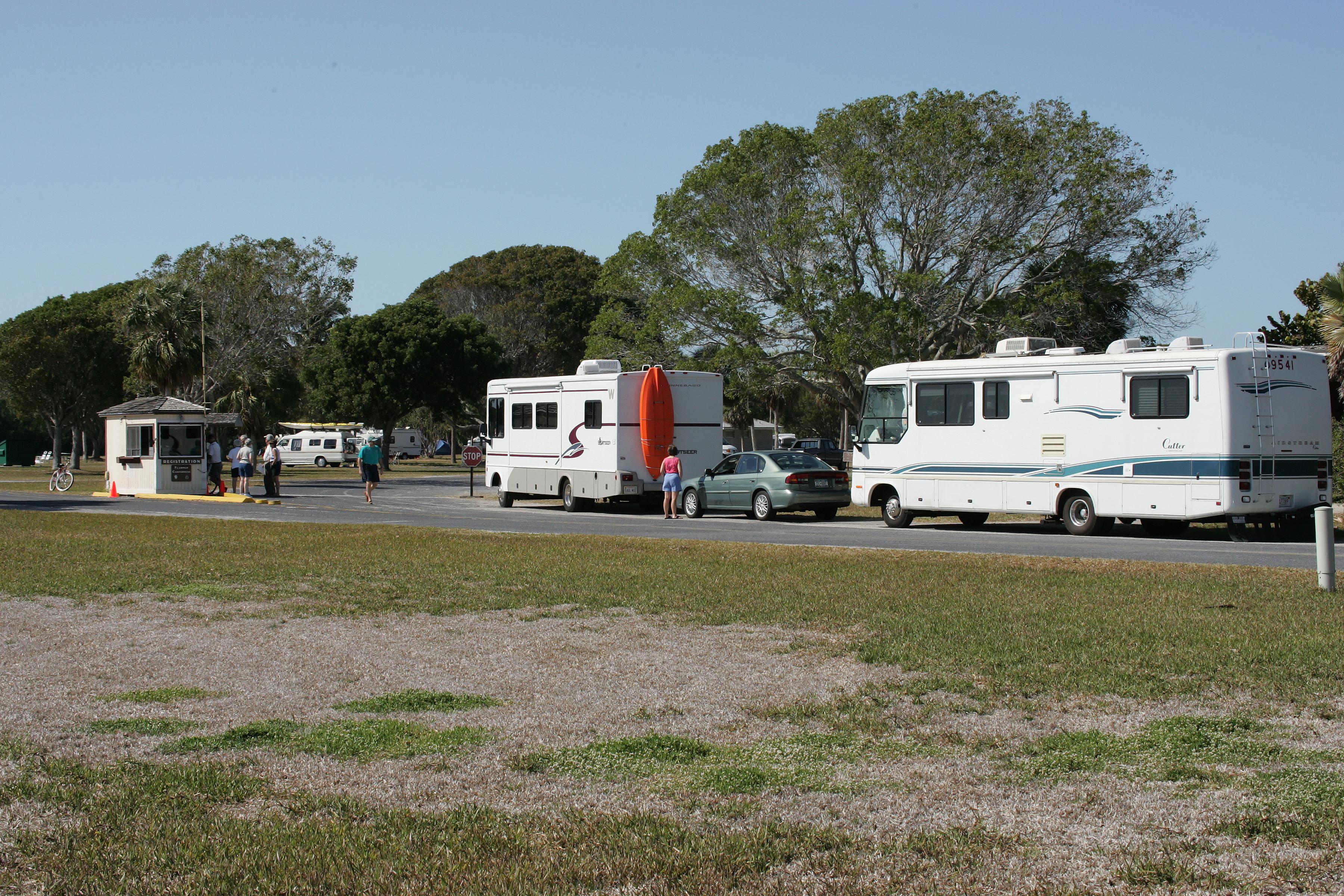 Two white recreational vehicles are in line to register for a campsite. Trees in the background.