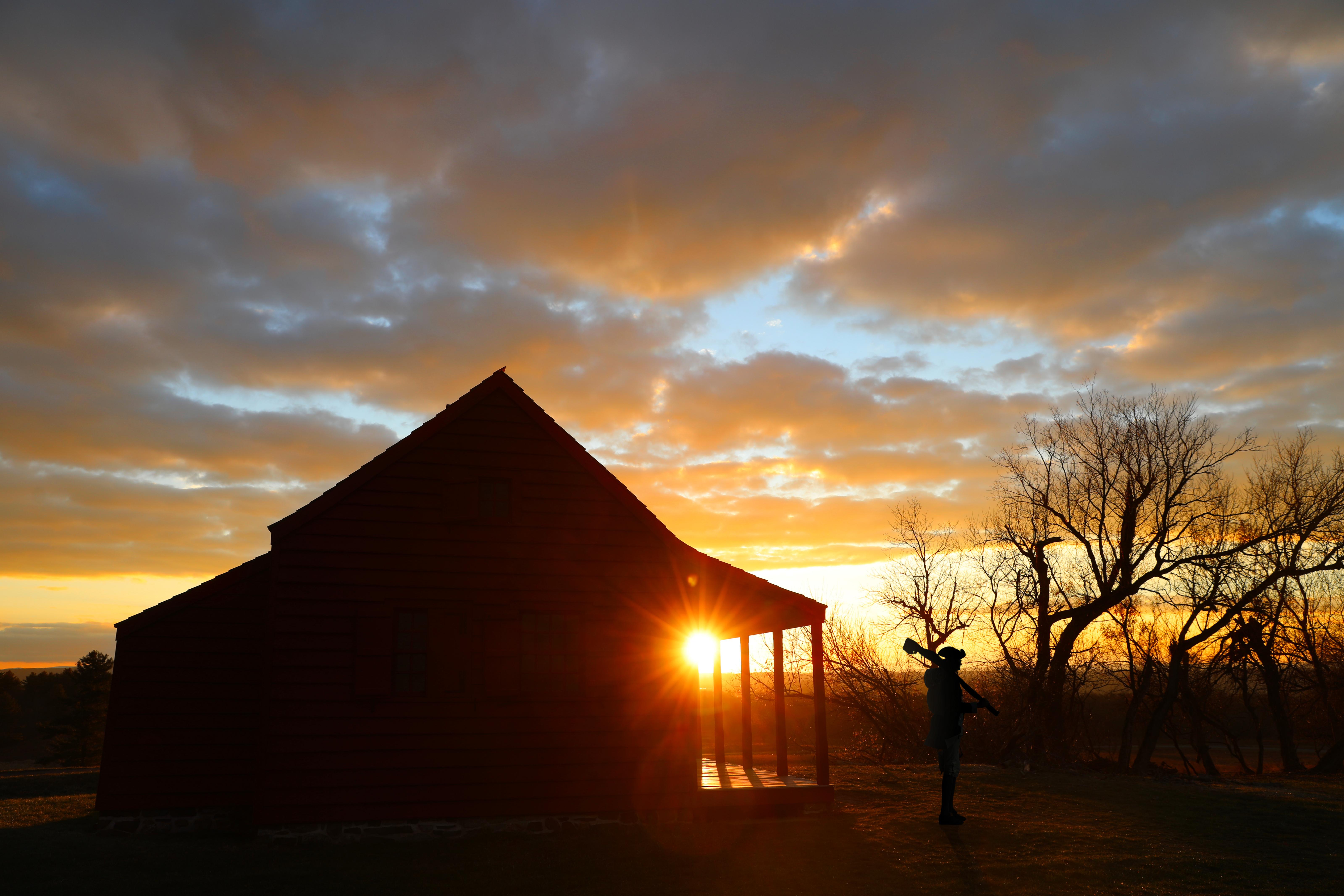 A man wearing 18th-century clothing carrying a musket and a farmhouse are silhouetted by sunrise.