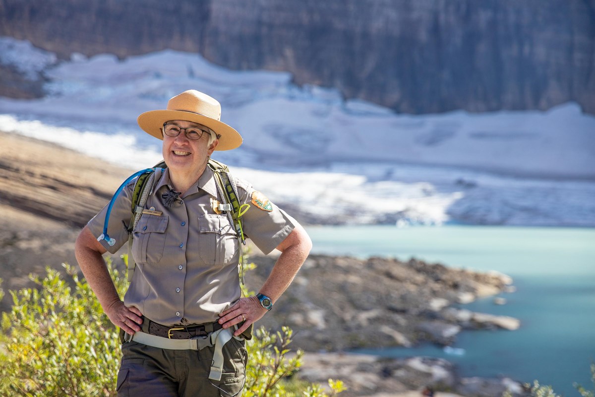 a park ranger in uniform stands in front of a glacier and a lake