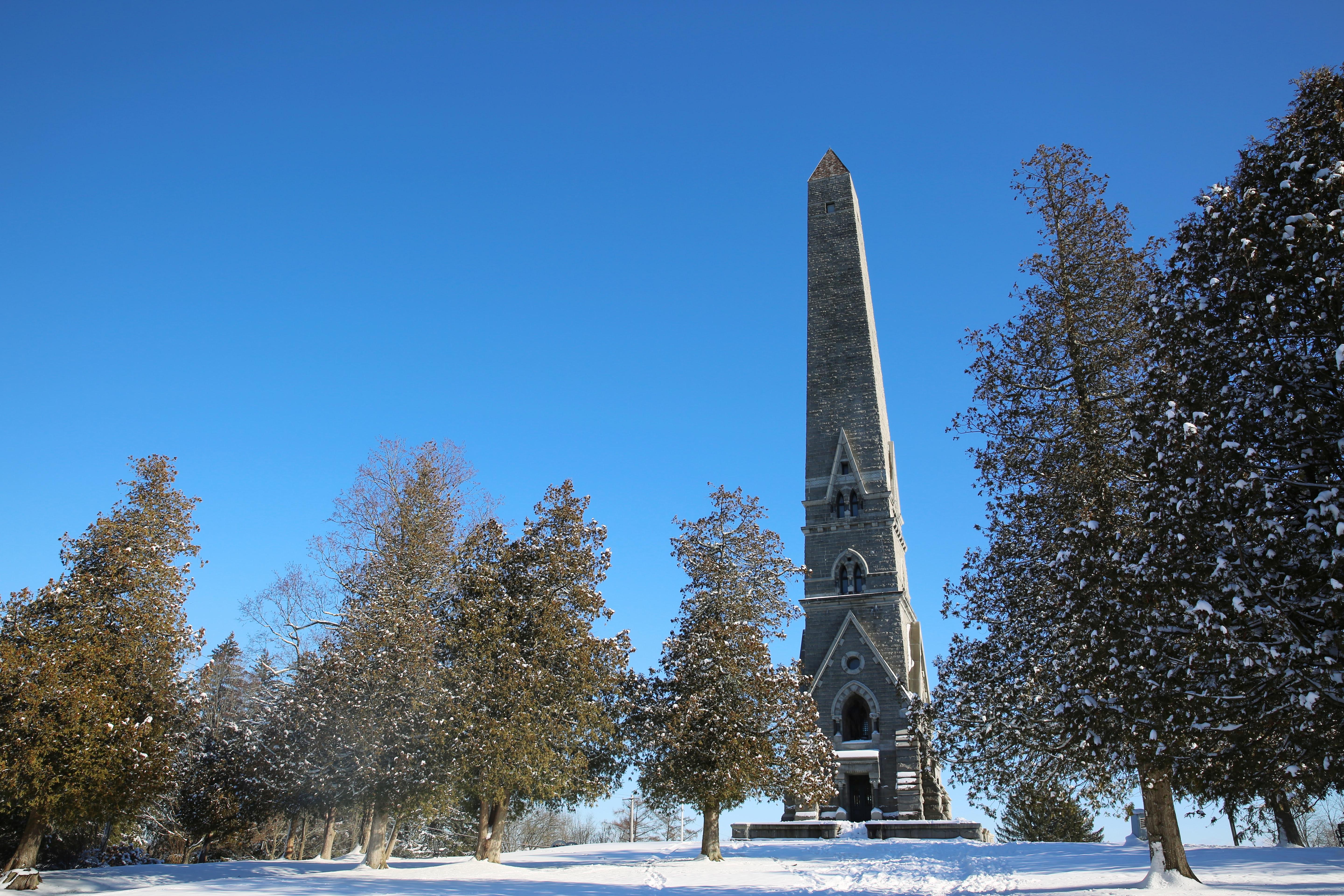 A narrow, stone obelisk on top of a snowy hill reaches into a clear sky.