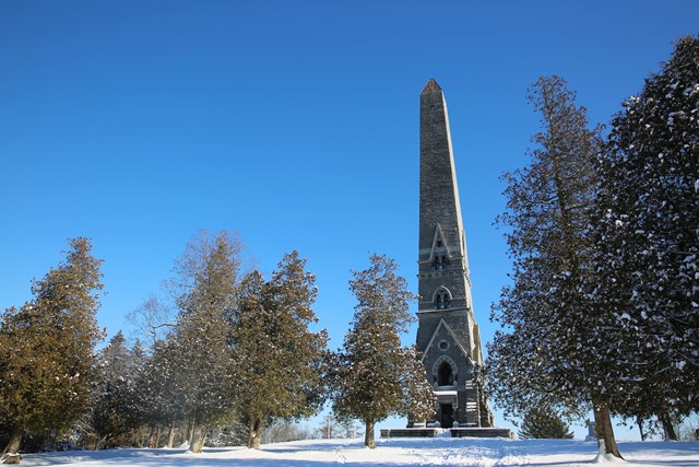 A narrow, stone obelisk on top of a snowy hill reaches into a clear sky.