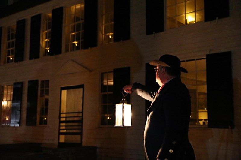 A man in 18th-century style clothing holds a candlelit lantern in front of a 2-story yellow house.