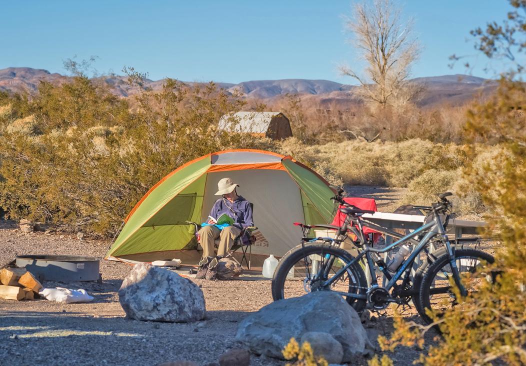 Camper sits on dirt ground outside tent with bicycles nearby.