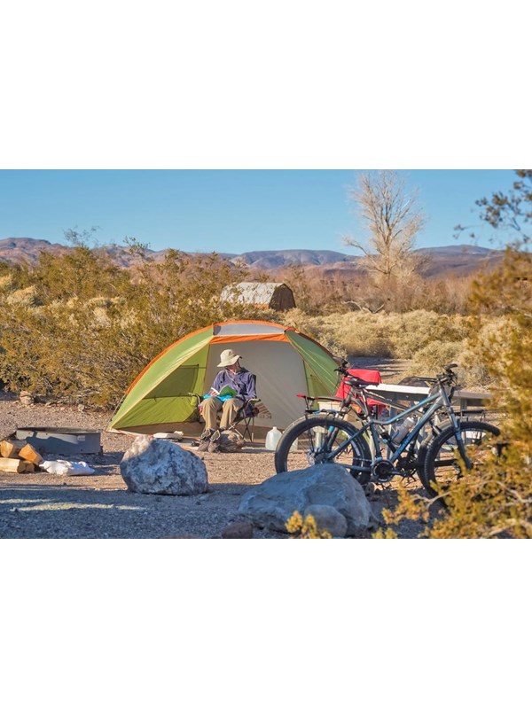 Camper sits on dirt ground outside tent with bicycles nearby.