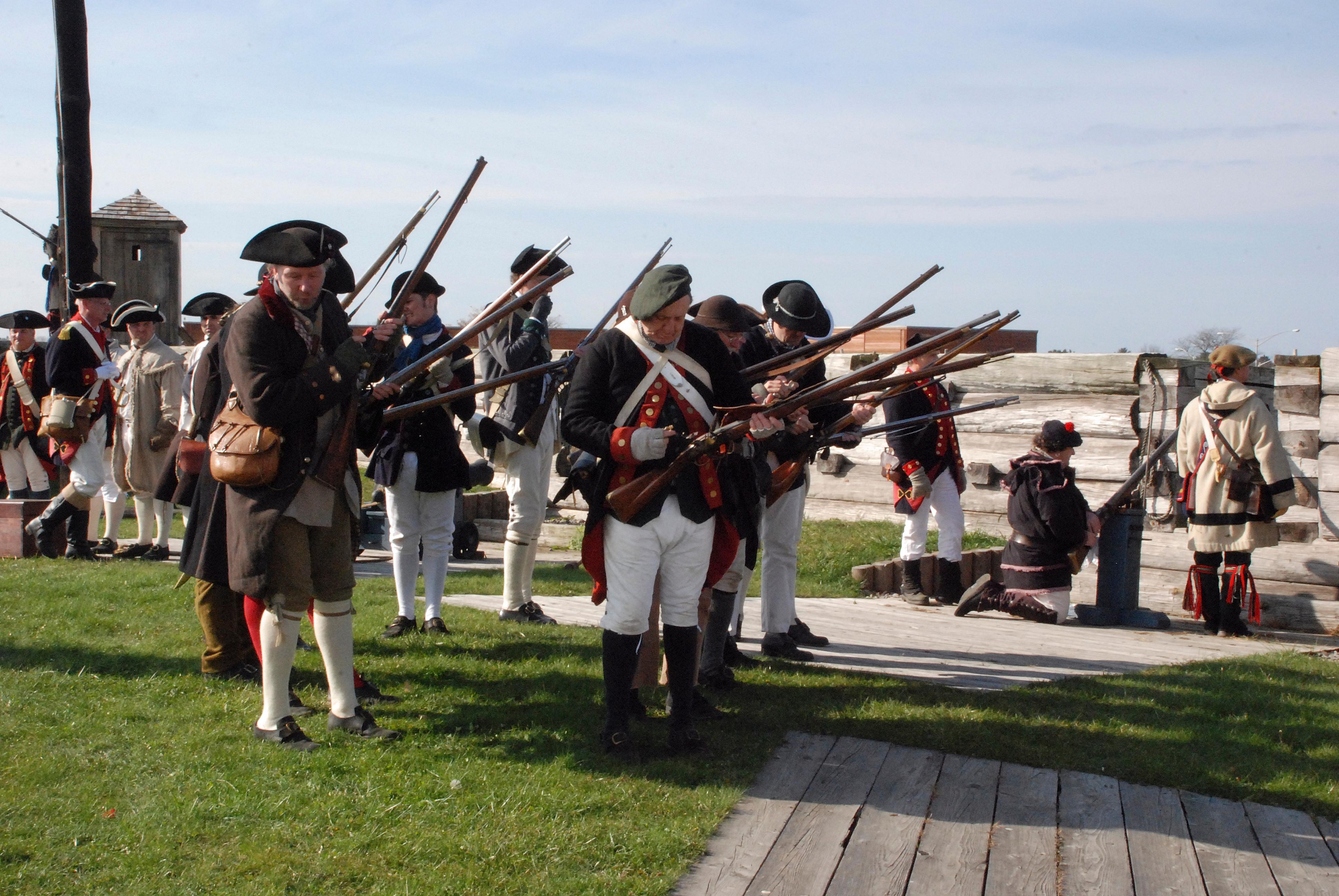 About two dozen soldiers stand in a neat rows holding their muskets.