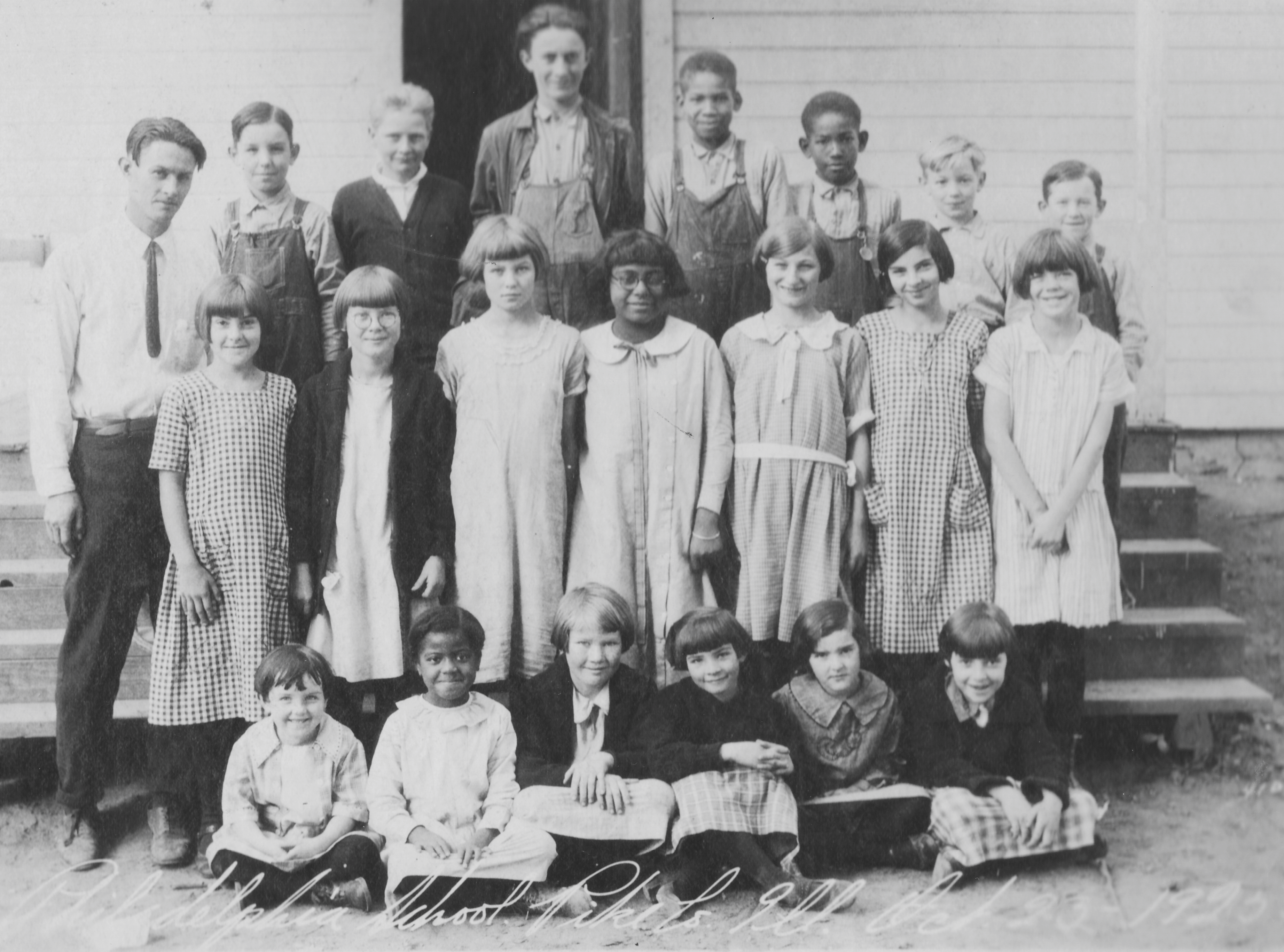 Twenty children of mixed ages and genders posing for the camera in front of a schoolhouse.