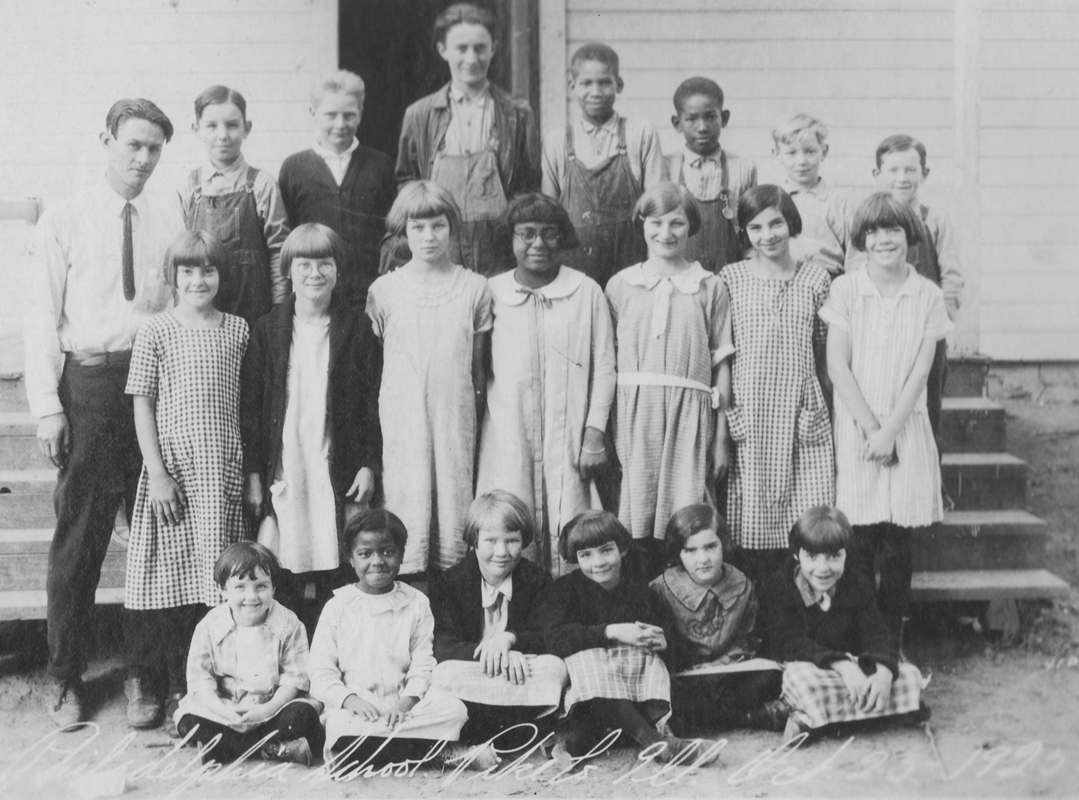 Twenty children of mixed ages and genders posing for the camera in front of a schoolhouse. 