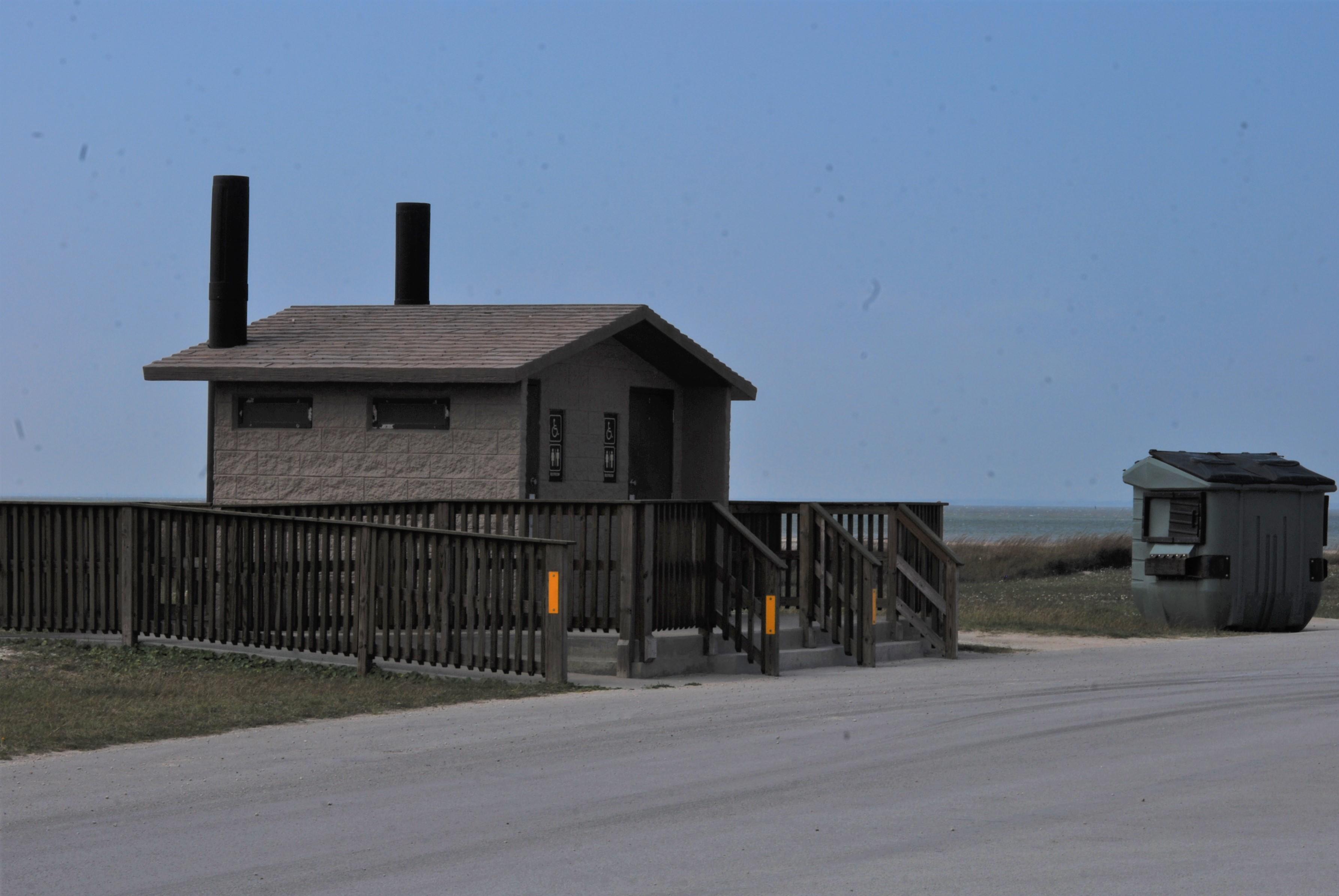 a brown wood fence outlines the ramp and stairs to a double vault toilet