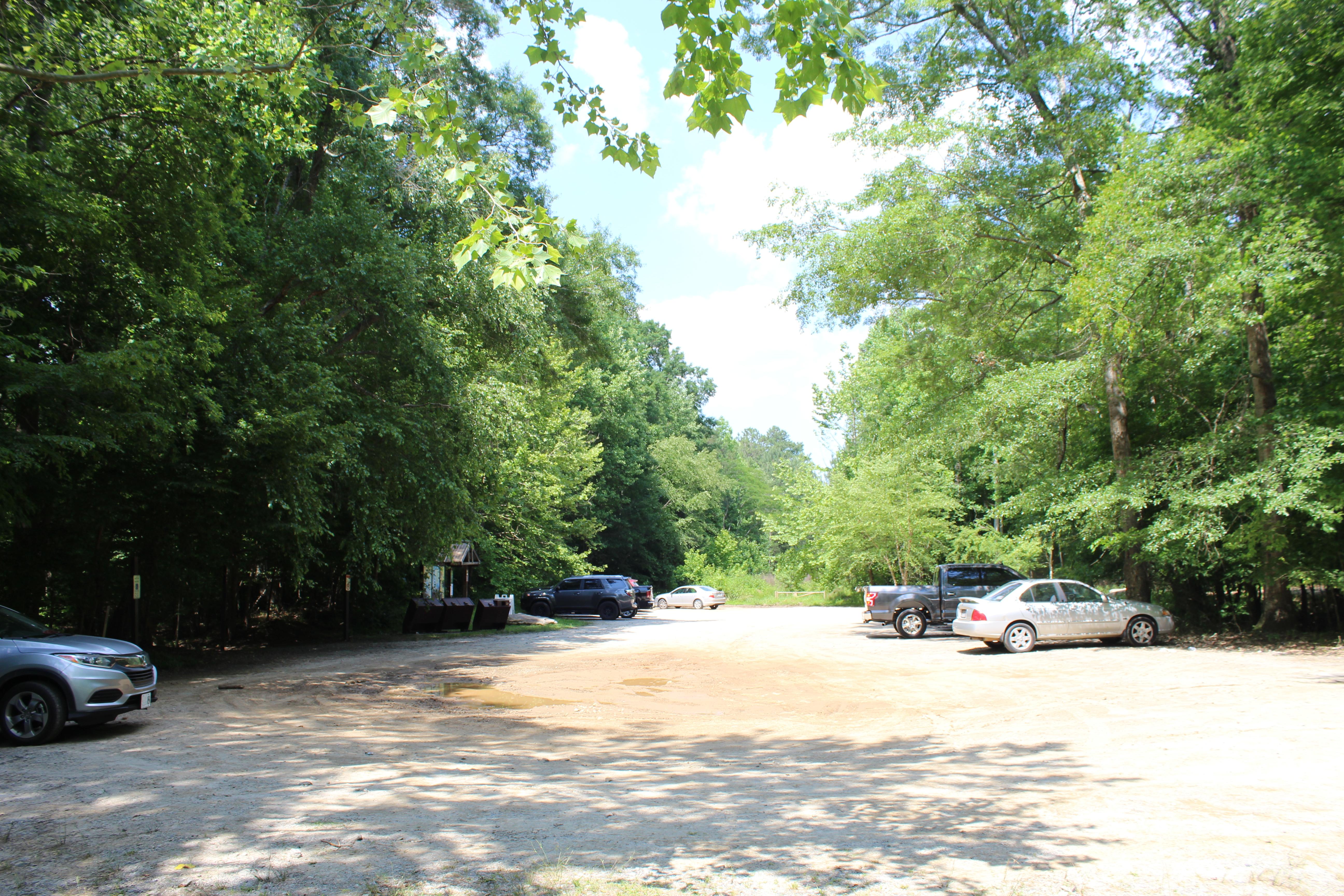 Gravel parking lot with several cars and trucks and mature trees along edge of lot.