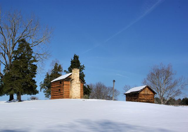 Back side of kitchen cabin and smokehouse in snow with blue sky and trees