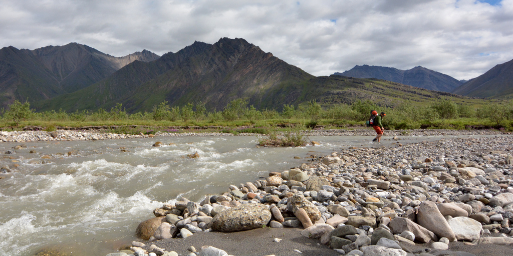 A hiker crosses a stream with mountains in the background