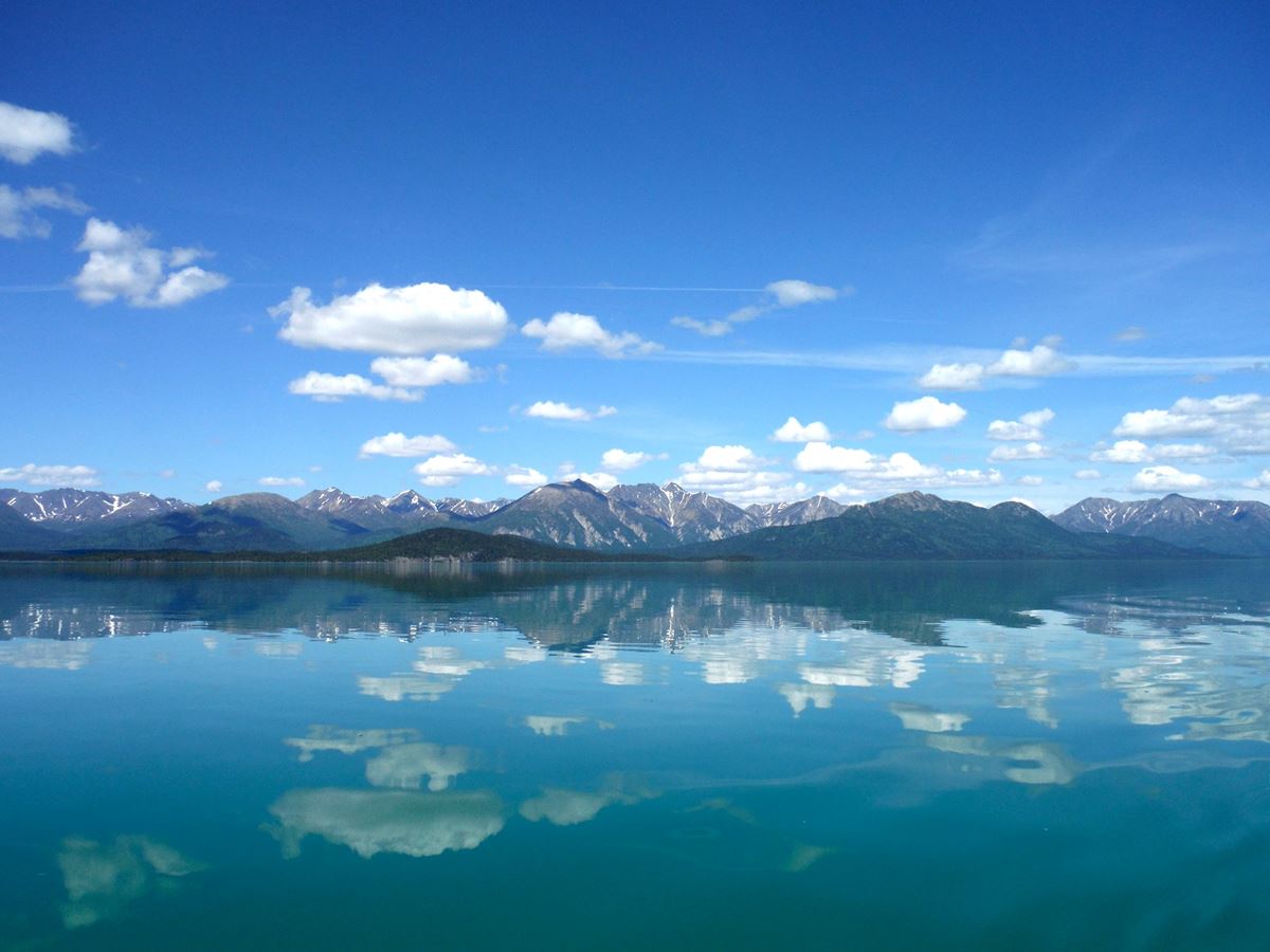 Photo of blue sky with fluffy white clouds reflect in calm lake with mountains in the background.