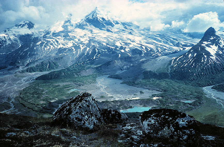 Photo of a stratovolcano flanked with glaciers towering over a river valley.