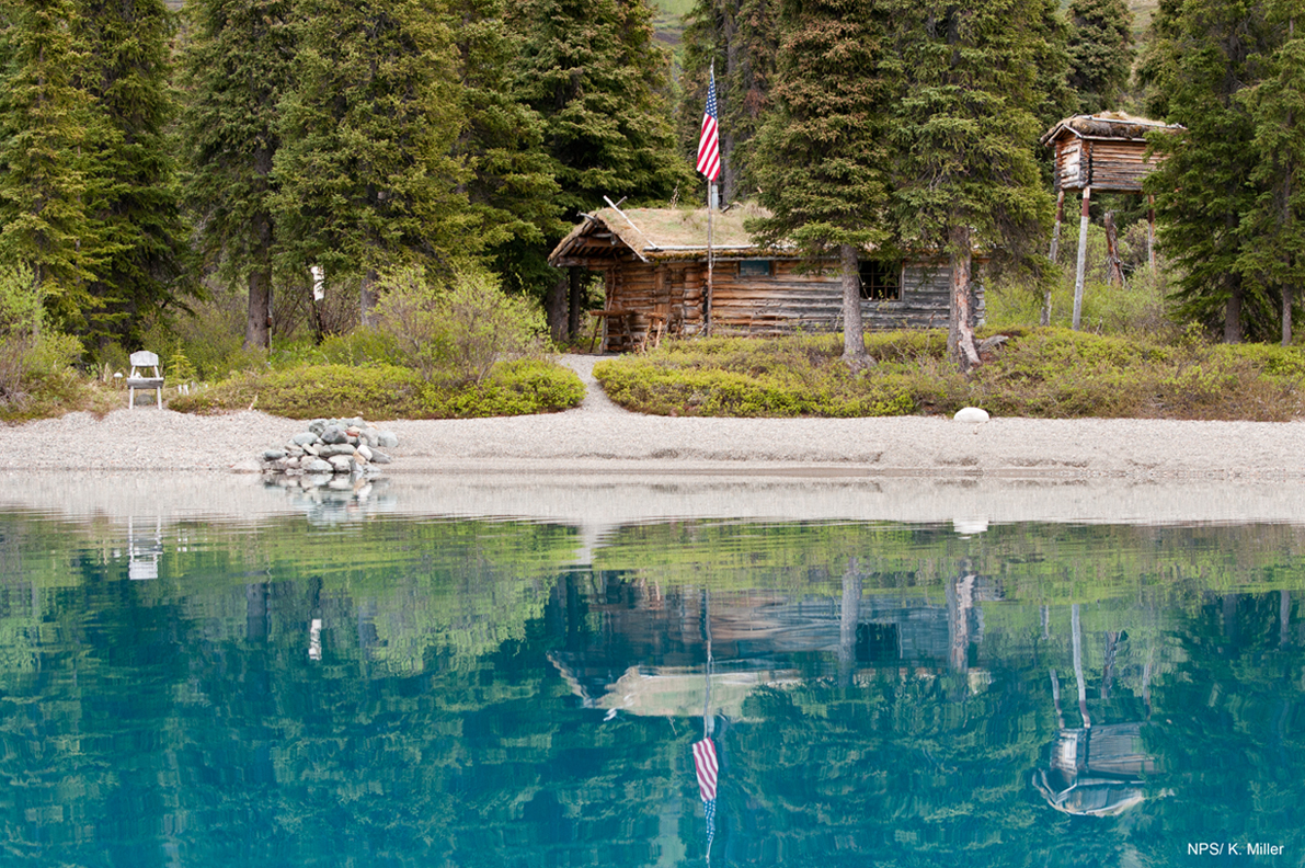 Photo of a one story log cabin, cache, forest, and American flag reflecting in a calm, blue lake.