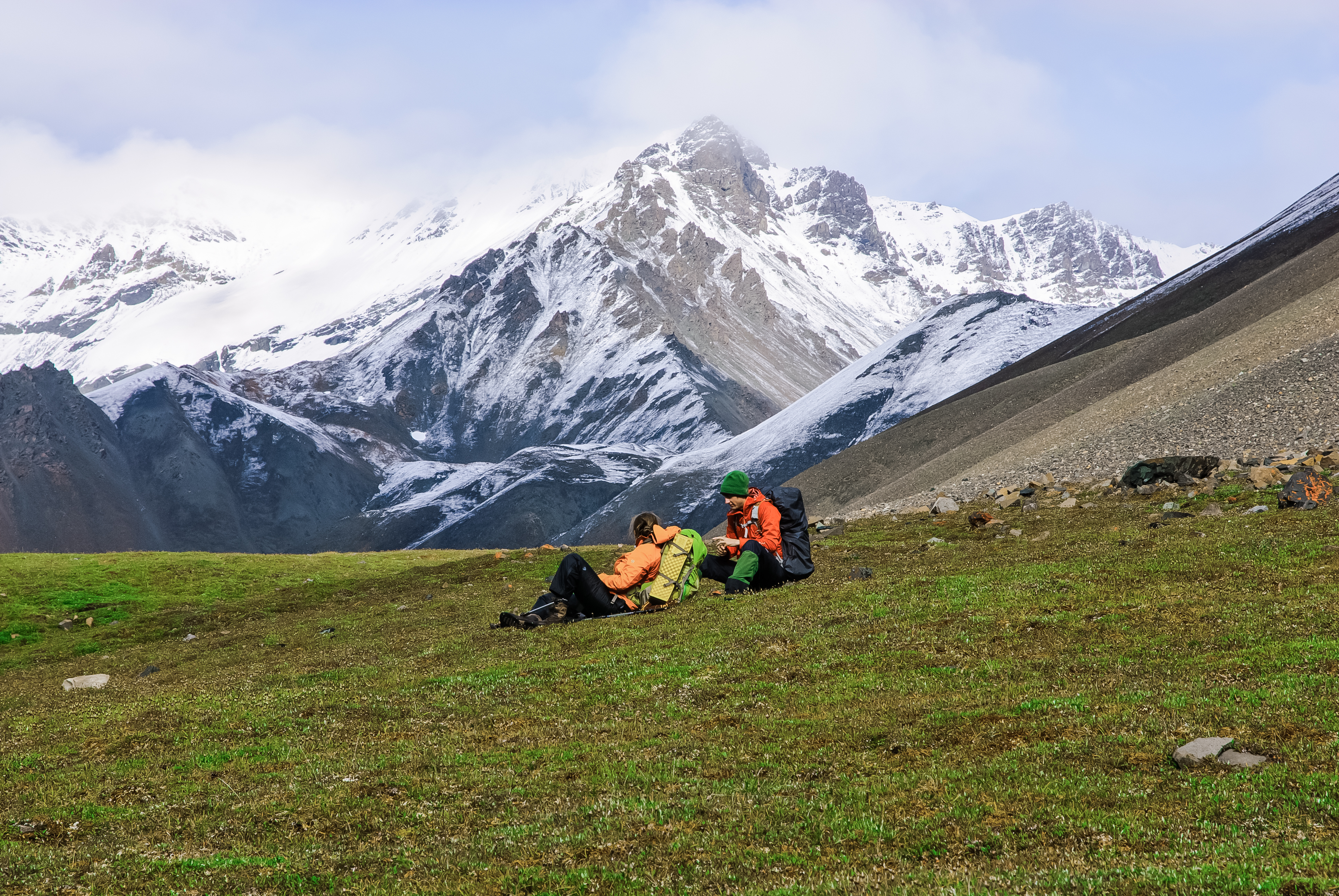 Two backpackers sitting in an alpine meadow with snowy mountains in the background