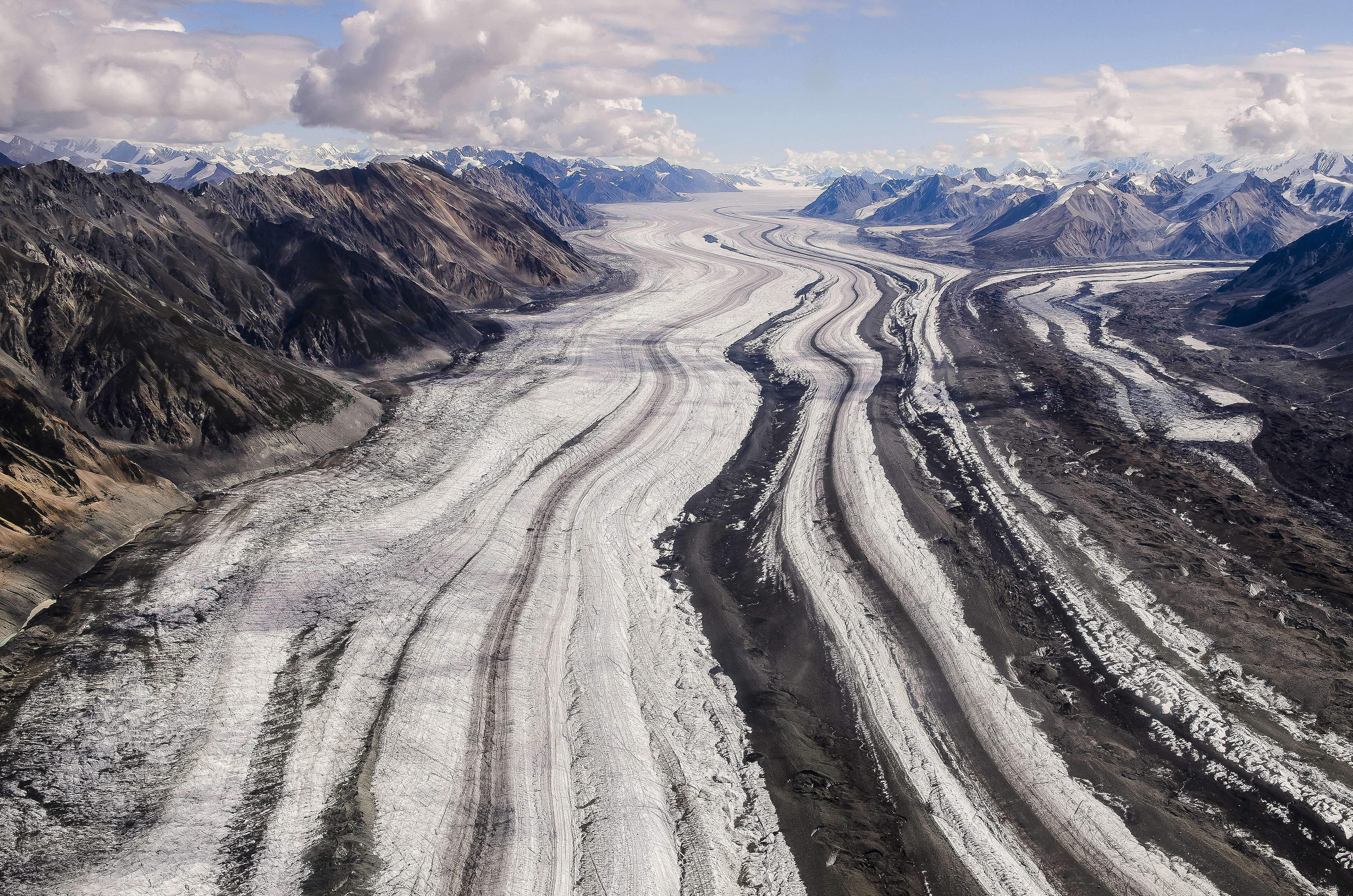 A large glacier with stripes of different colored rock nestled in between barren mountain slopes.