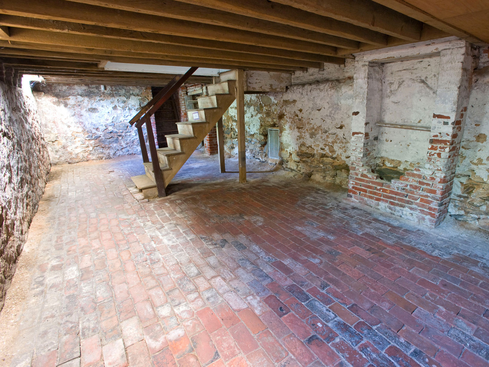 Color photo of the basement with brick floor, false chimney, and wooden staircase.