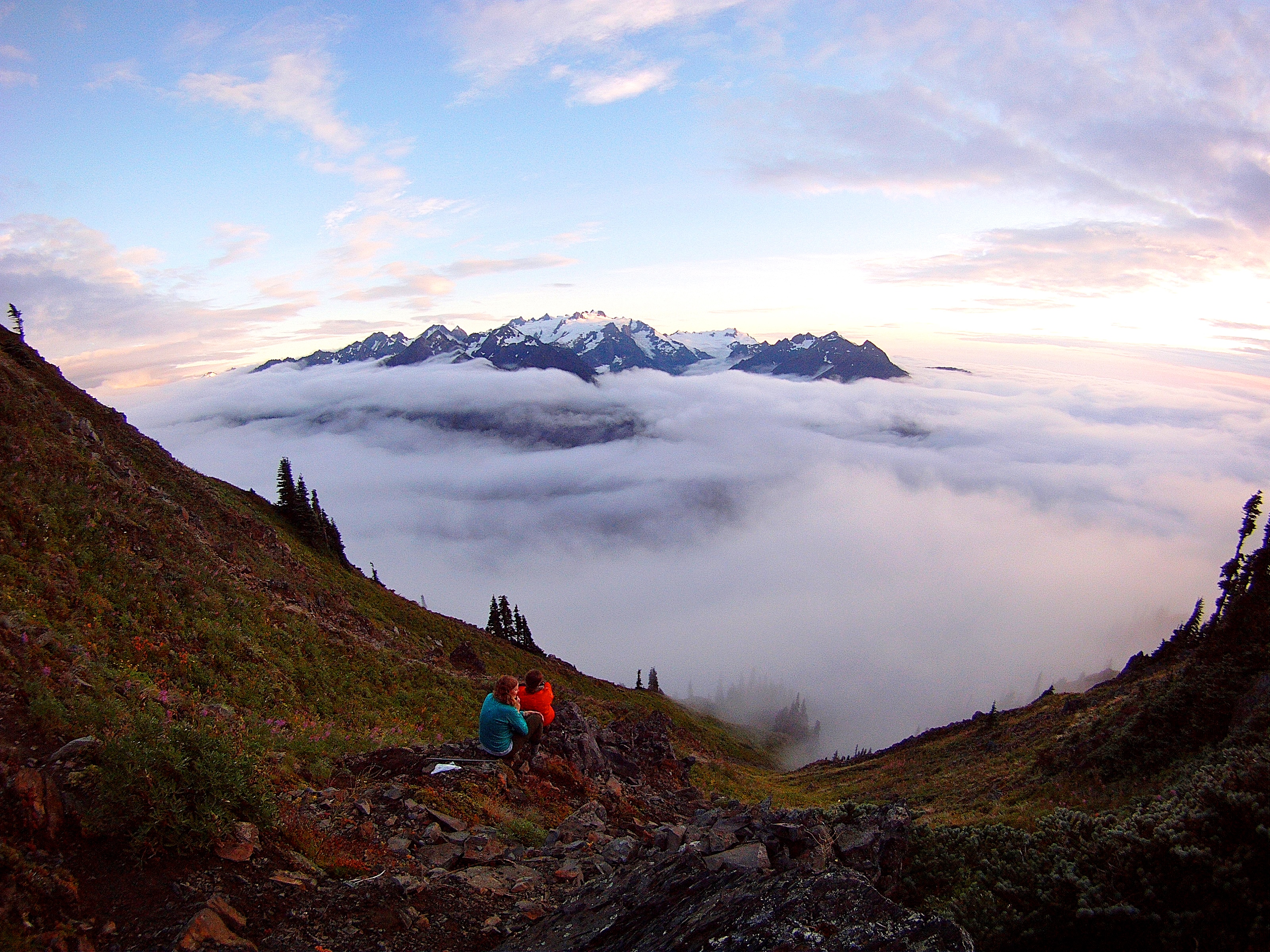 Hikers sit and watch the sun set behind snow-capped mountains.