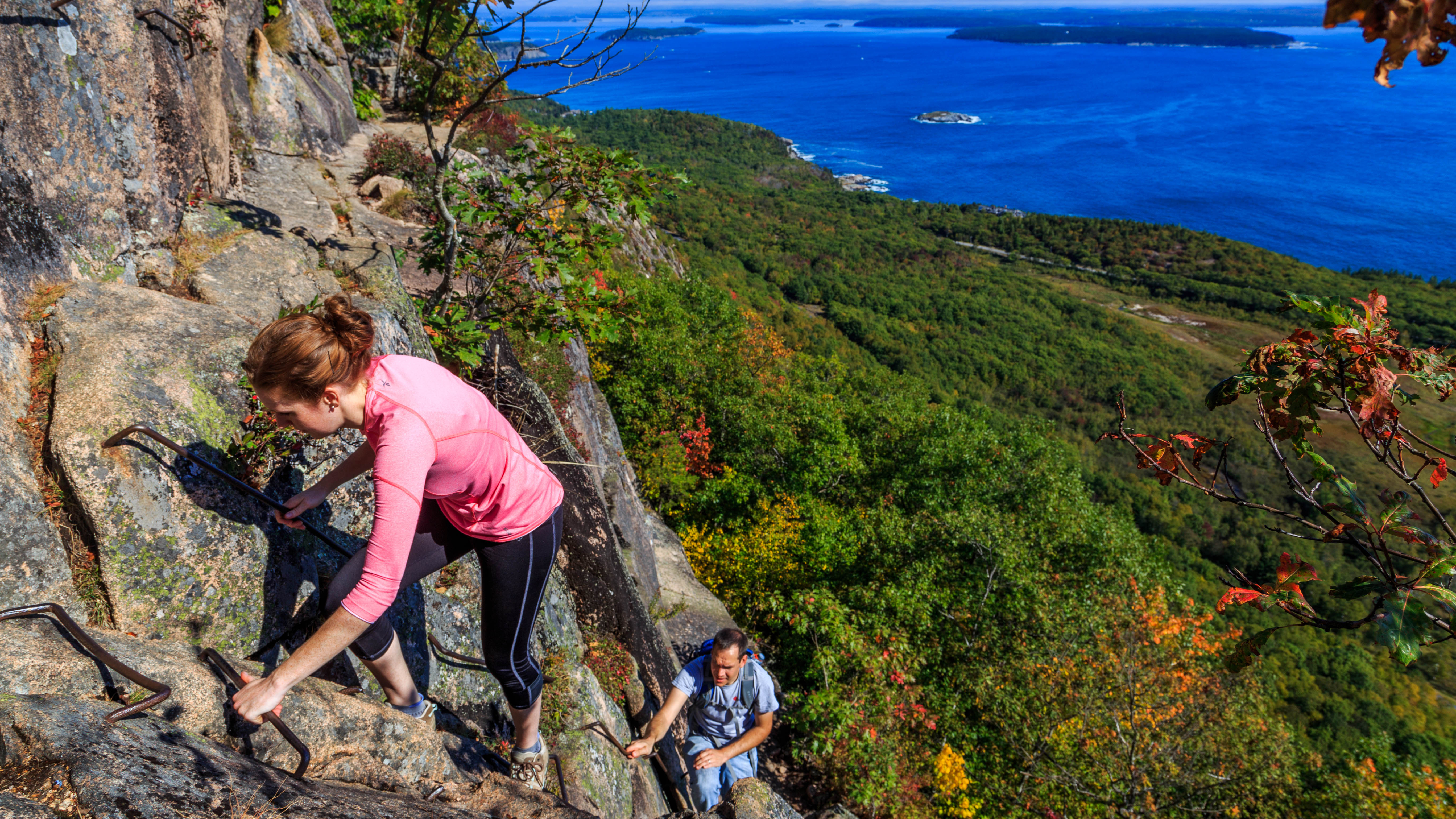 Two hikers ascend a sheer cliff trail by way of historic iron rung ladders.