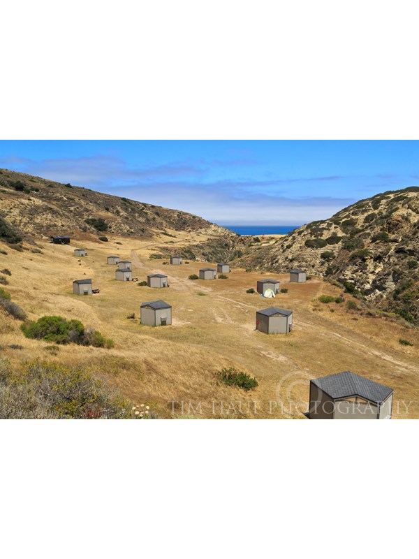 Campgrounnd wind shelters on dry, grassy terrace looking down canyon to blue ocean.